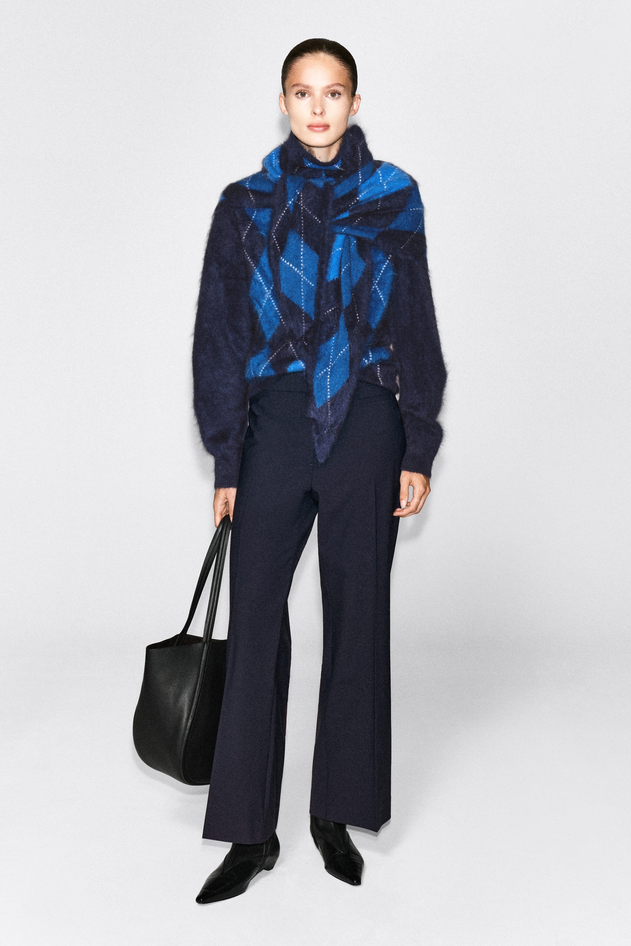 An argyle brushed cashmere triangle scarf in shades of blue and navy is worn wrapped around the neck and draped over the model’s chest. The scarf is styled with a matching patterned jumper, dark wide-leg trousers, and a black leather tote bag carried in one hand. An argyle brushed cashmere triangle scarf in shades of blue and navy is worn wrapped around the neck and draped over the model’s chest. The scarf is styled with a matching patterned jumper, dark wide-leg trousers, and a black leather tote bag carried in one hand.