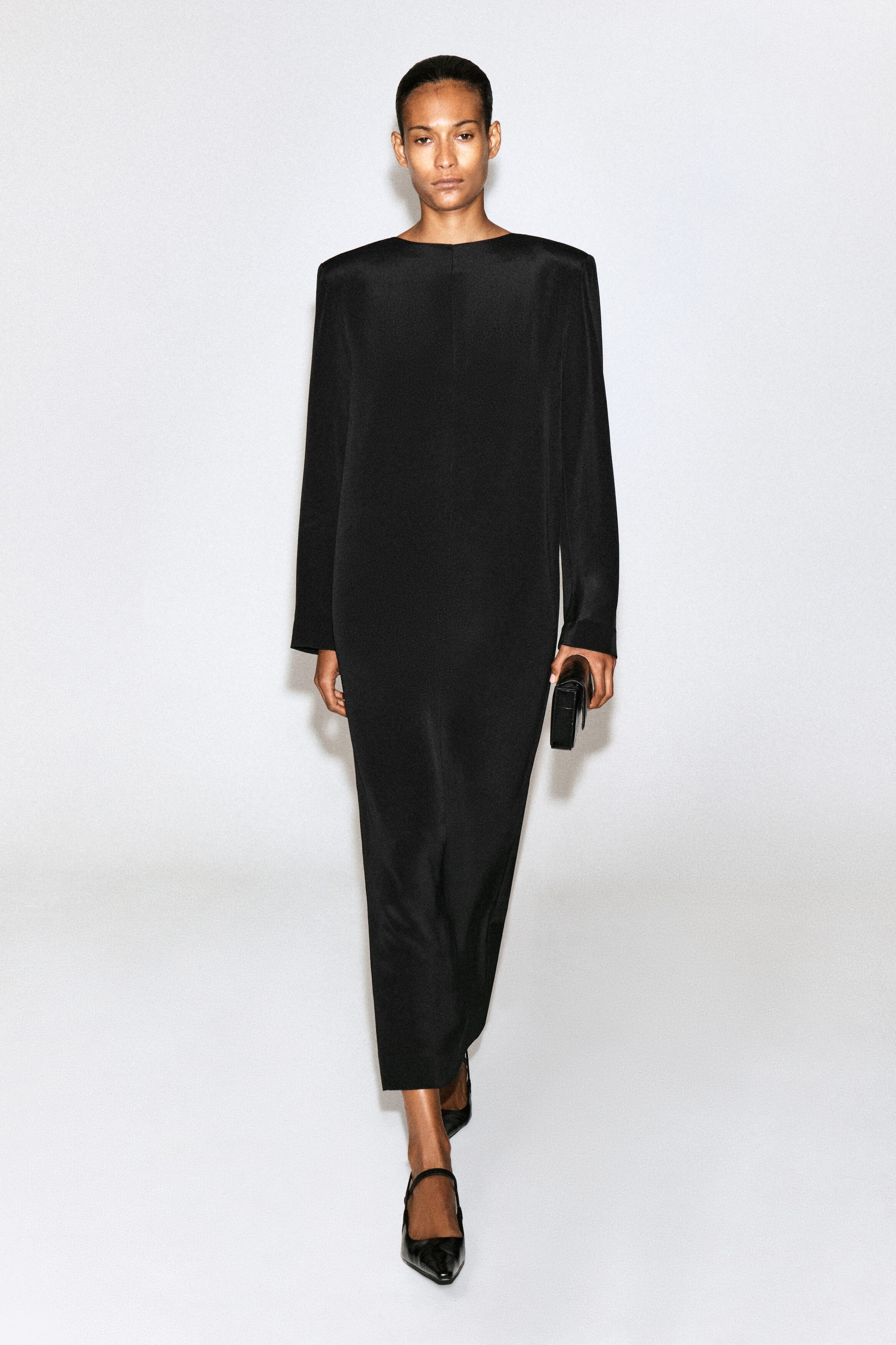 A black maxi dress featuring structured shoulders, long sleeves, and a straight silhouette is being worn by the model. The model is holding a small black clutch in one hand while walking forwards.