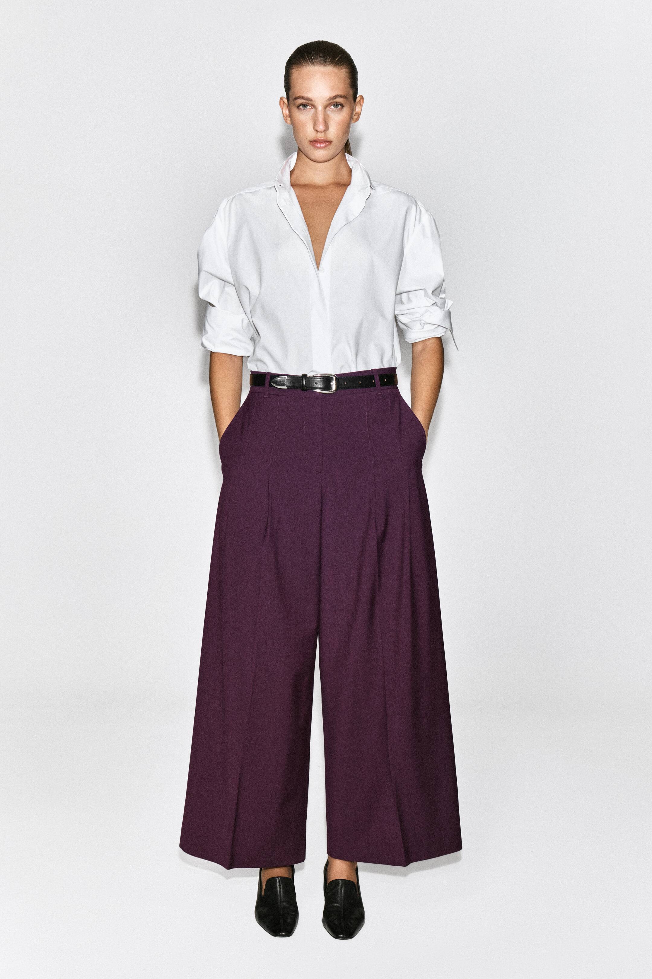 Agrandir l'image: Un pantalon large en mélange de laine plissé, couleur prune foncée, est porté par le mannequin, qui se tient debout avec les mains dans les poches latérales. Ce pantalon taille haute présente des plis sur le devant et est associé à une chemise blanche à col rentrée à l’intérieur, une ceinture en cuir noire et des chaussures plates noires.