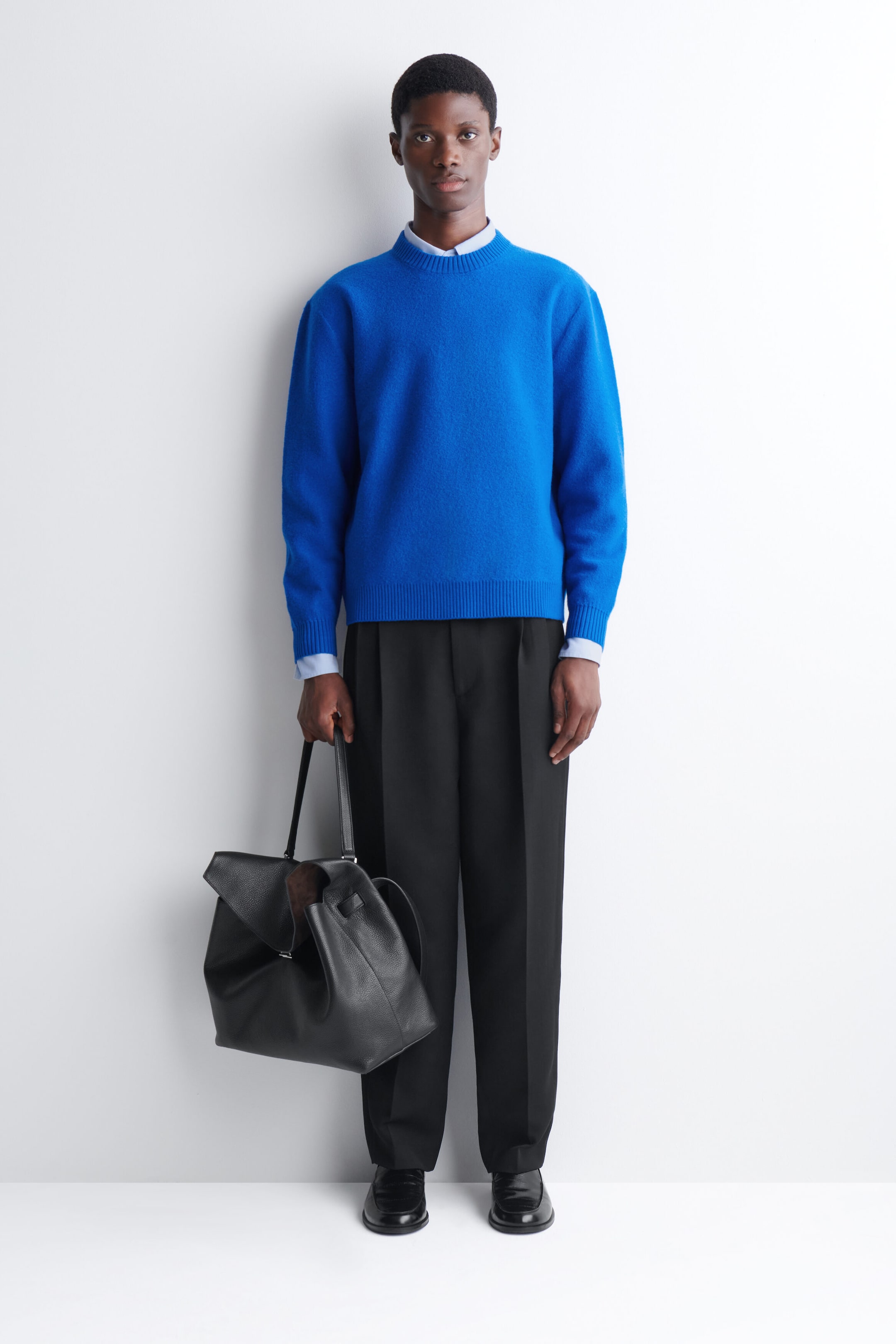 A bright blue knitted crew-neck sweater with ribbed trims is worn by the model over a light blue collared shirt. The sweater is styled with black trousers, and the model is holding a black leather bag. A bright blue knitted crew-neck sweater with ribbed trims is worn by the model over a light blue collared shirt. The sweater is styled with black trousers, and the model is holding a black leather bag.
