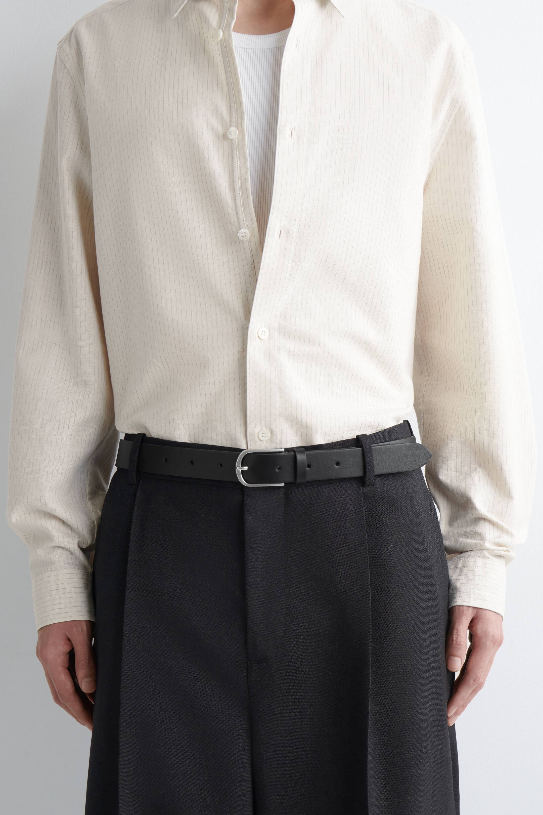 Преглед на по-голямо изображение: Черeн кожен колан със сребриста тока е носен от модела. Той пристяга черен плисиран панталон, комбиниран със светла риза на райета.