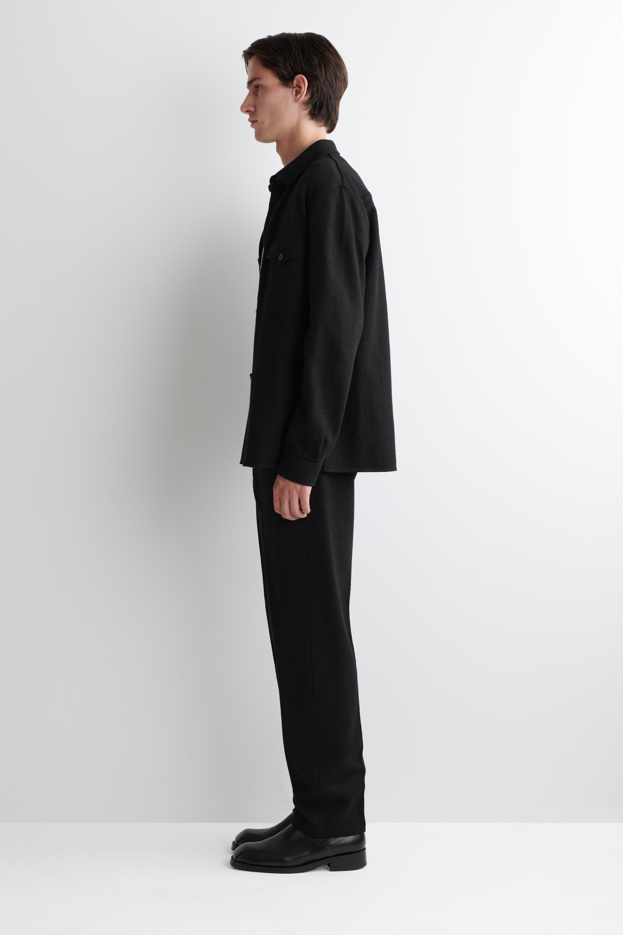 Agrandir l'image: Une surchemise noire pour homme et un pantalon assorti sont présentés de côté gauche. La surchemise présente un col classique, des manches longues, une poche poitrine visible et une coupe décontractée et carrée. Le pantalon noir assorti est conçu avec une jambe droite et une coupe décontractée, et est porté avec des chaussures noires simples.