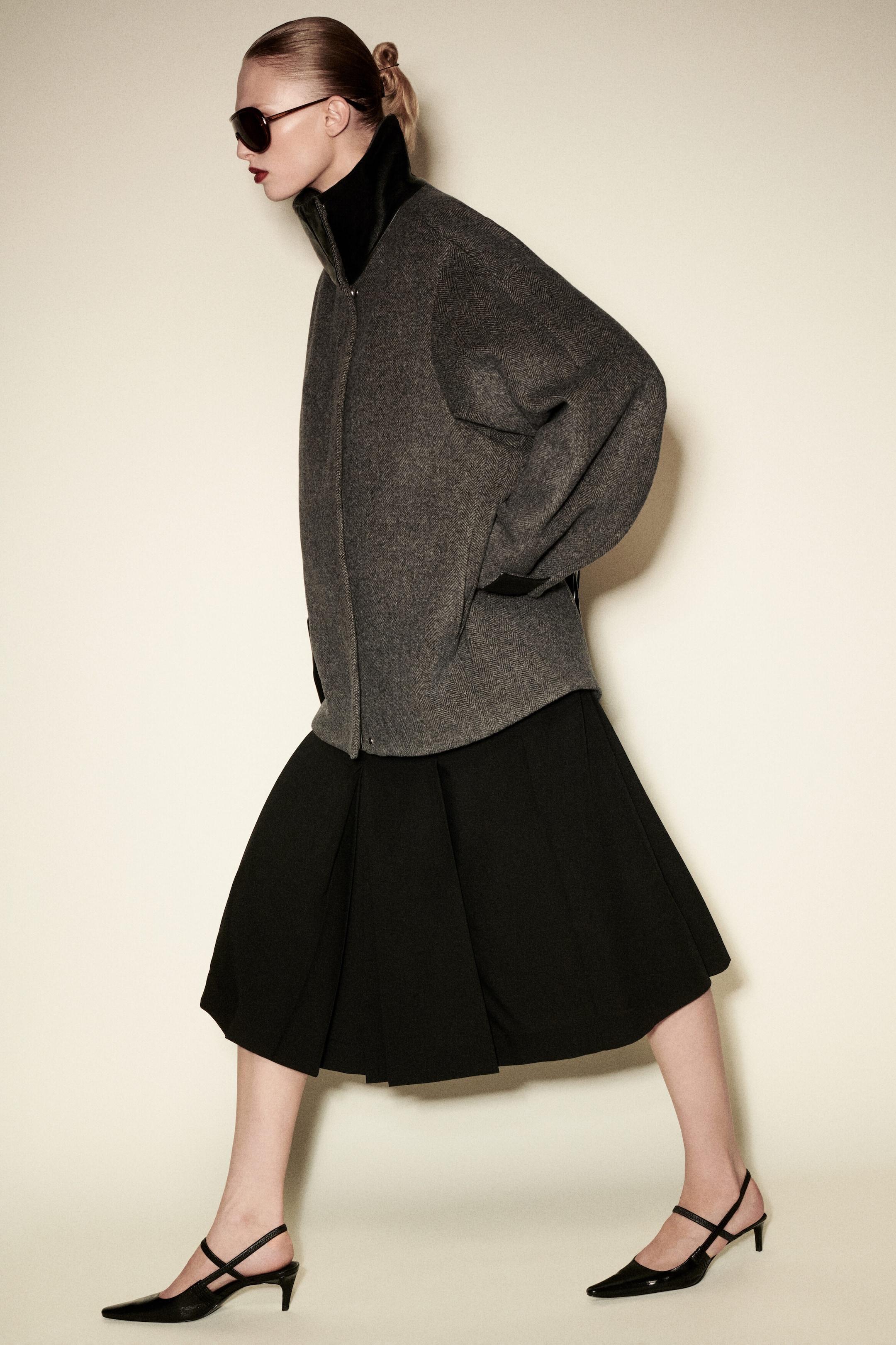 Eine graue Jacke aus Wollmischung im Fischgrätmuster zeigt sich in einer seitlichen Profilansicht mit Oversize-Silhouette und einem hohen schwarzen Funnelkragen aus Leder. Die Jacke ist mit voluminösen Ärmeln, schwarzen verstellbaren Manschettenriegeln und einem geschwungenen Saum gestaltet. Eine graue Jacke aus Wollmischung im Fischgrätmuster zeigt sich in einer seitlichen Profilansicht mit Oversize-Silhouette und einem hohen schwarzen Funnelkragen aus Leder. Die Jacke ist mit voluminösen Ärmeln, schwarzen verstellbaren Manschettenriegeln und einem geschwungenen Saum gestaltet.