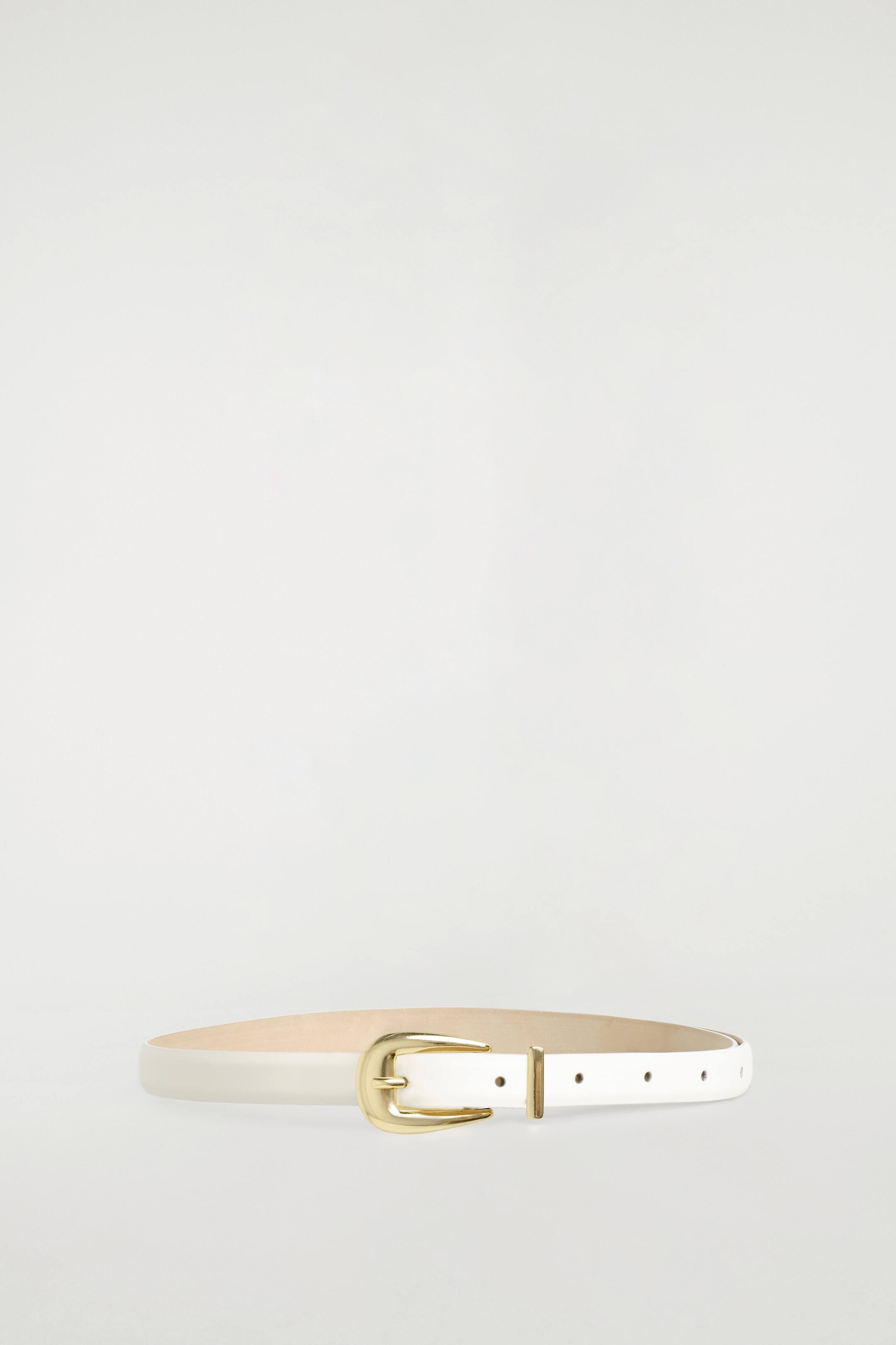 Agrandir l'image: La ceinture en cuir à boucle sculptée repose en boucle fermée, face à l’avant, mettant en valeur sa lanière en cuir blanc lisse avec une doublure ton naturel. Elle présente une boucle en métal doré poli au profil courbé, accompagnée d’un passant en métal assorti et de trous de réglage pré-perforés.