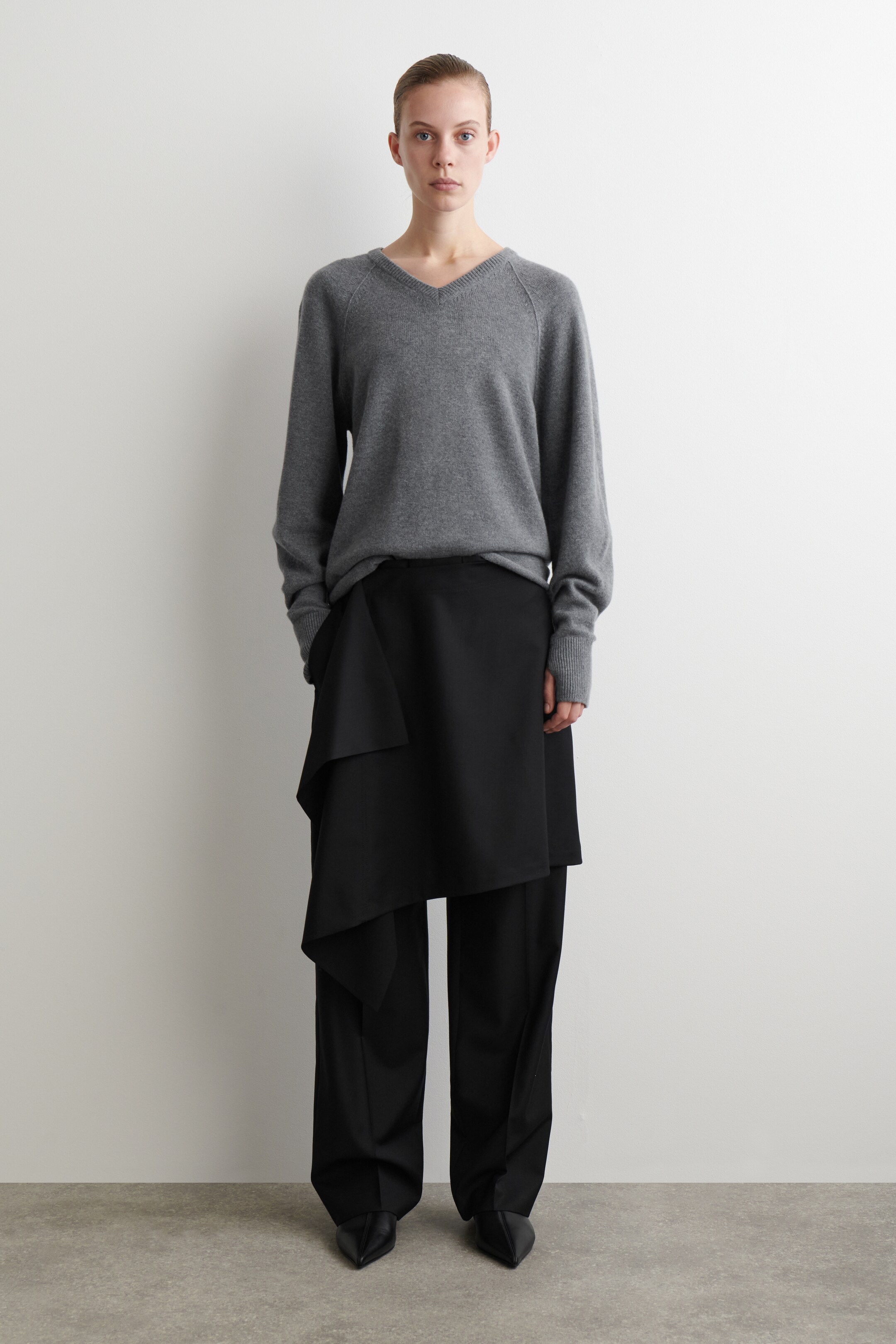 Agrandir l'image: Un pull gris à col en V avec des manches raglan et des poignets côtelés est porté, présentant une coupe décontractée. Il est superposé à un pantalon noir à jambes droites, partiellement recouvert par une superposition asymétrique de style enveloppant noir, avec des ballerines noires à bout pointu complétant l'ensemble.