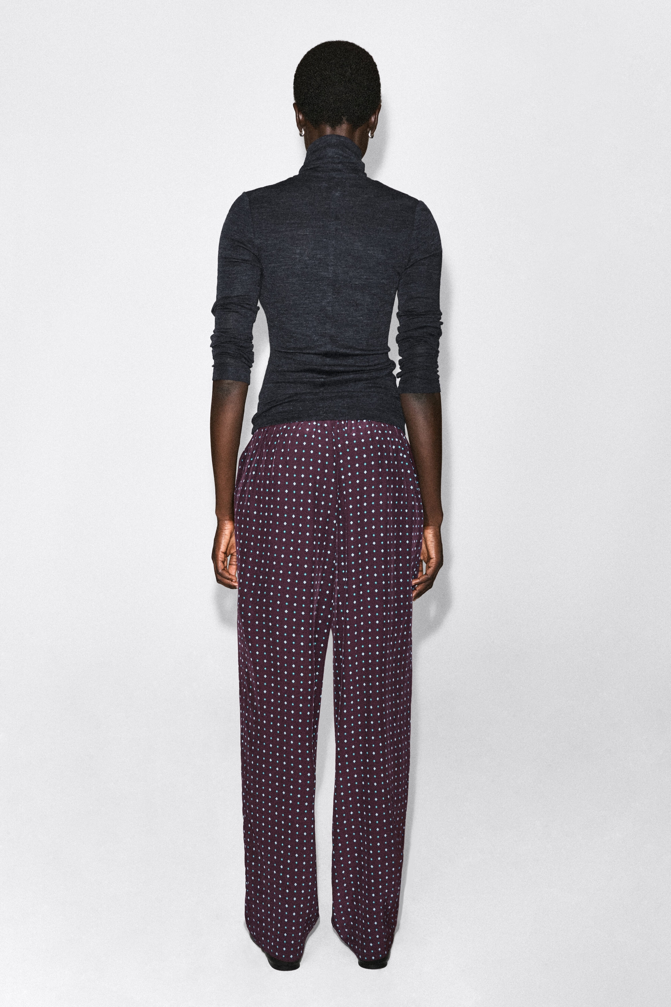 Le pantalon droit à motif losanges est présenté de dos, avec une coupe décontractée dans un tissu violet foncé orné d’un motif losange bleu clair sur l’ensemble. Le pantalon tombe droit jusqu’au sol et est associé à un haut à col roulé à manches longues gris foncé.