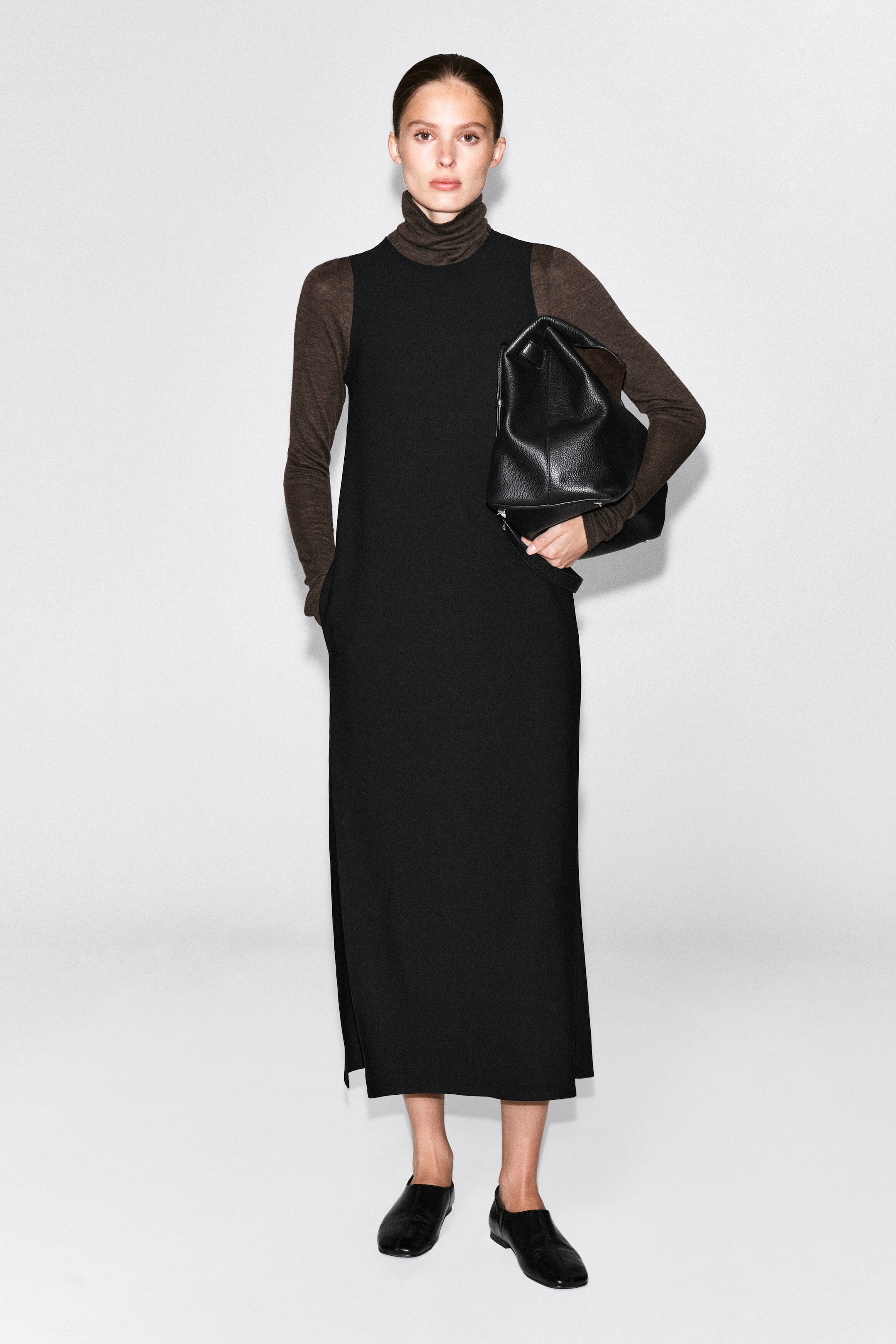 The model is wearing a black sleeveless crepe midi dress layered over a long-sleeve brown turtleneck top. The dress features a straight silhouette, side pockets, and a side-slit hem, with the model placing one hand in a pocket while holding a large black leather bag under their arm.