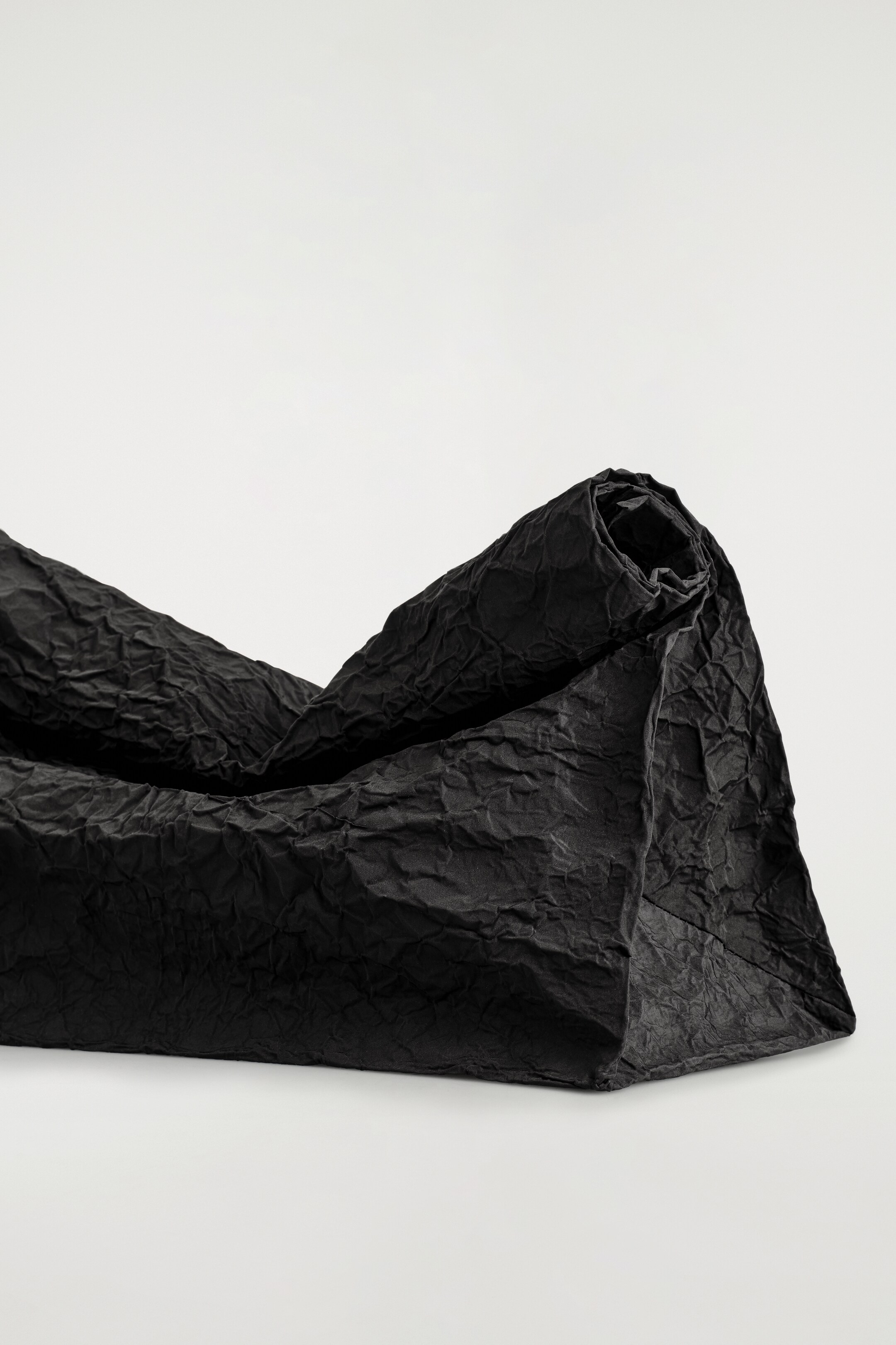Grösseres Bild ansehen: Eine schwarze Clutch-Tasche liegt horizontal und zeigt ihre zerknitterte Textur und übergroße Silhouette. Der obere Teil der Tasche ist nach innen gerollt und bildet einen kompakten Verschluss, während der flache Boden sichtbar ist und eine stabile Basis bietet.