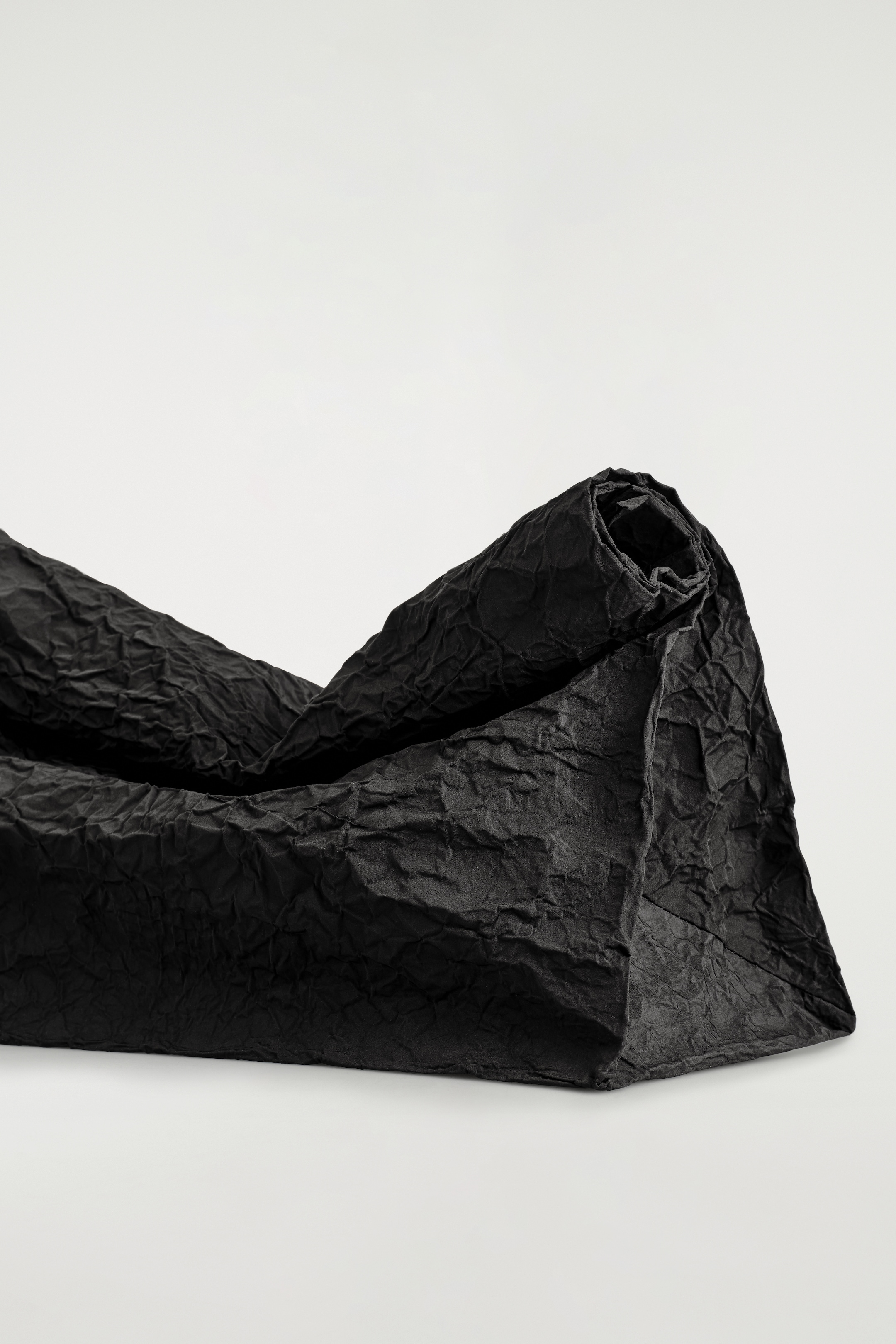 Visualizza immagine più grande: Una pochette nera è posizionata orizzontalmente, mostrando la sua texture increspata e la silhouette oversize. La parte superiore della borsa è arrotolata verso l'interno, formando una chiusura compatta, mentre il suo fondo piatto è visibile, fornendo una base stabile.