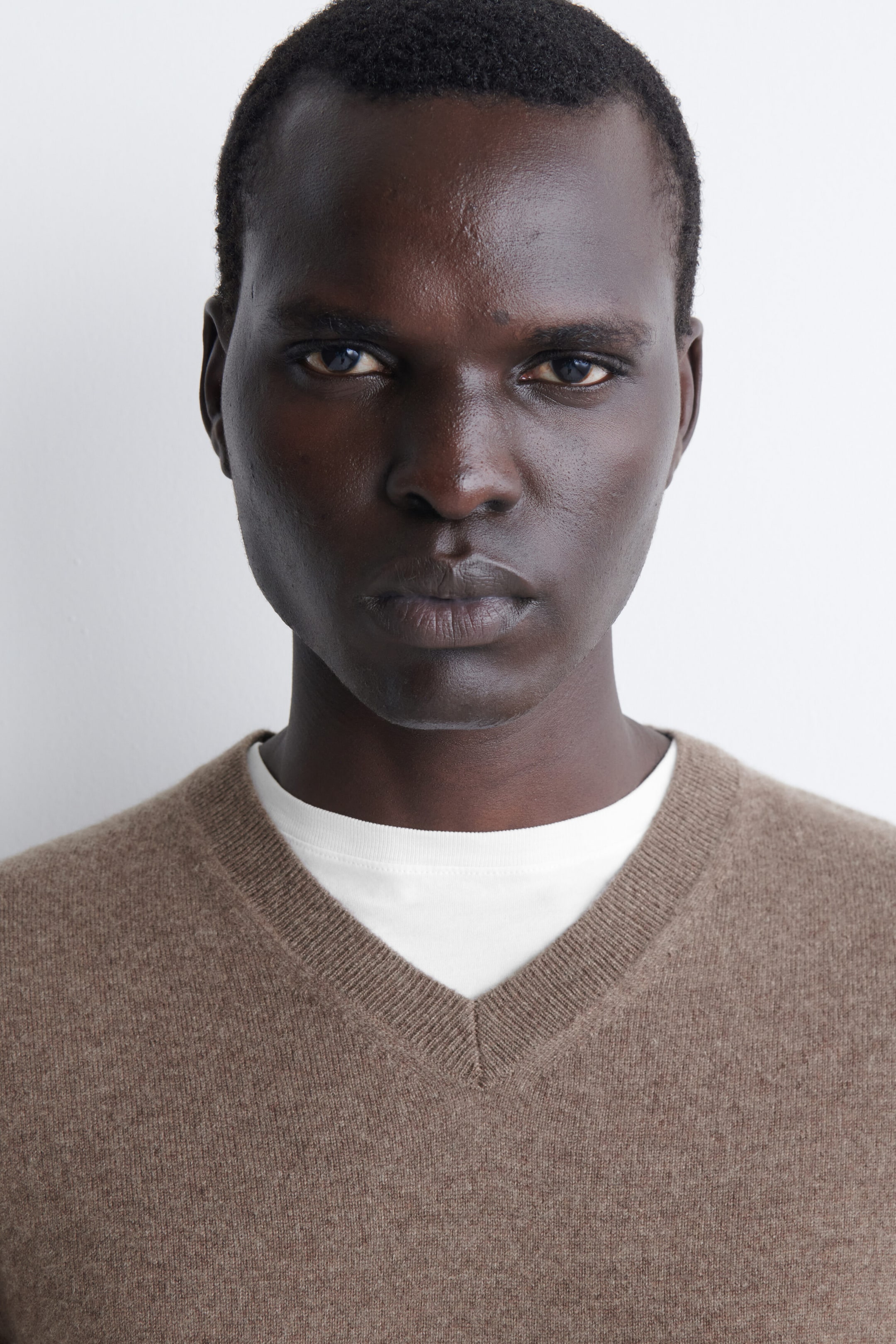 The cashmere V-neck jumper in a light brown mélange is shown in a front-facing close-up, highlighting its ribbed collar and soft, fine-knit texture. The jumper is being worn layered over a white crew-neck T-shirt, emphasising its classic fit and neckline shape. The cashmere V-neck jumper in a light brown mélange is shown in a front-facing close-up, highlighting its ribbed collar and soft, fine-knit texture. The jumper is being worn layered over a white crew-neck T-shirt, emphasising its classic fit and neckline shape.