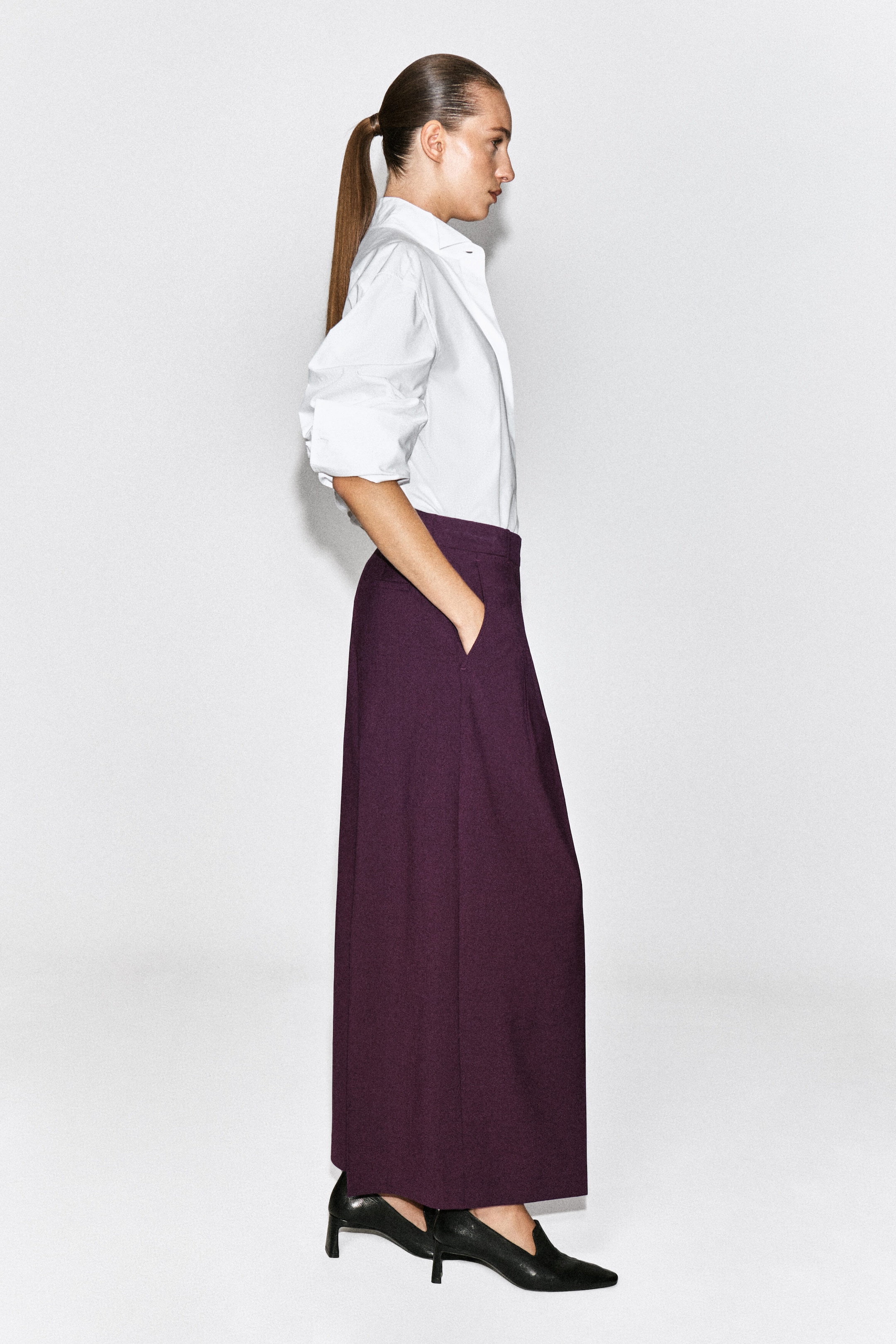 Pantaloni ampi plissettati in misto lana, color prugna intenso, indossati dalla modella, che è in piedi di profilo con una mano appoggiata nella tasca laterale. I pantaloni a vita alta sono abbinati a una camicia bianca abbottonata infilata dentro e a scarpe nere con tacco e punta affusolata. Pantaloni ampi plissettati in misto lana, color prugna intenso, indossati dalla modella, che è in piedi di profilo con una mano appoggiata nella tasca laterale. I pantaloni a vita alta sono abbinati a una camicia bianca abbottonata infilata dentro e a scarpe nere con tacco e punta affusolata.