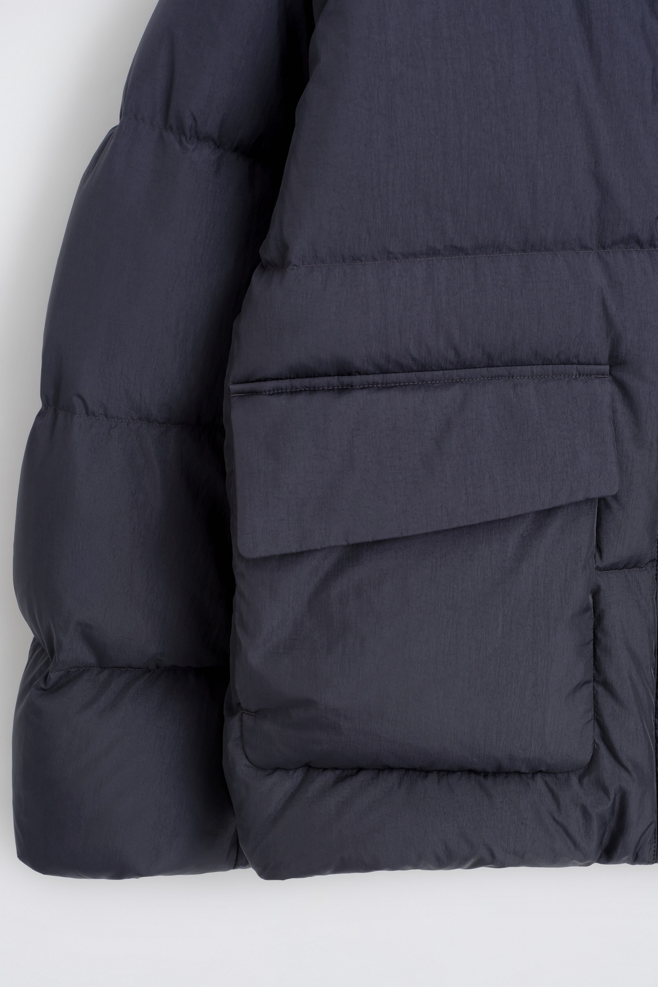 The funnel-neck nylon down puffer jacket is shown in a close-up view of its dark navy textured fabric, horizontal quilting, and large front flap pocket. The voluminous, down-filled puffer sections define the structure of the body and sleeves.