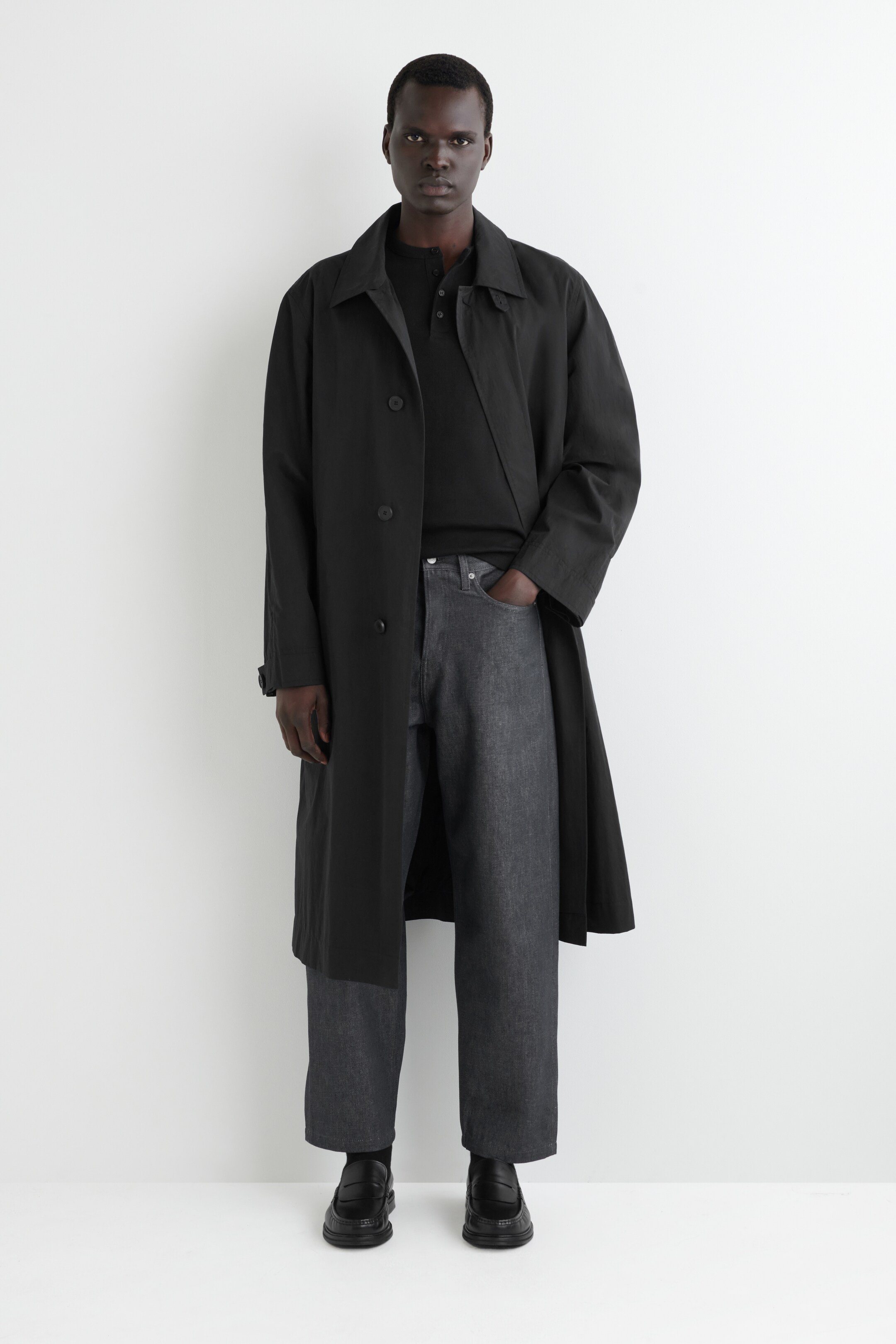 View larger image: A full outfit is being presented on a person facing forward, featuring a black, relaxed-fit trench coat with a classic collar and visible front buttons. This coat is being worn over a black long-sleeved polo shirt and paired with dark grey wide-leg trousers and black chunky loafers.