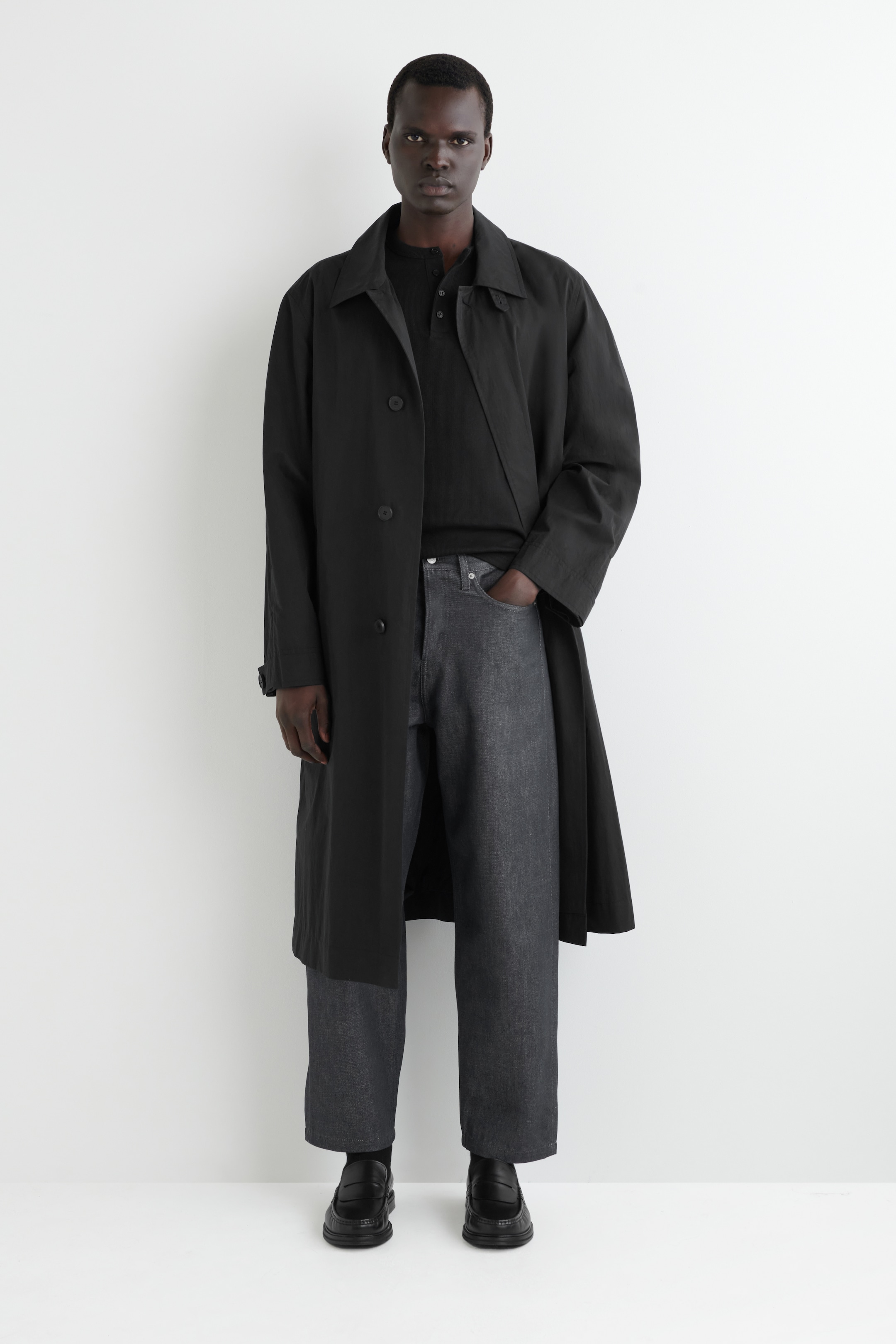 Agrandir l'image: Une tenue complète est présentée sur une personne faisant face à l'avant, avec un trench-coat noir à coupe ample, doté d'un col classique et de boutons visibles à l'avant. Ce manteau est porté par-dessus un polo noir à manches longues et associé à un pantalon large gris foncé et des mocassins noirs épais.