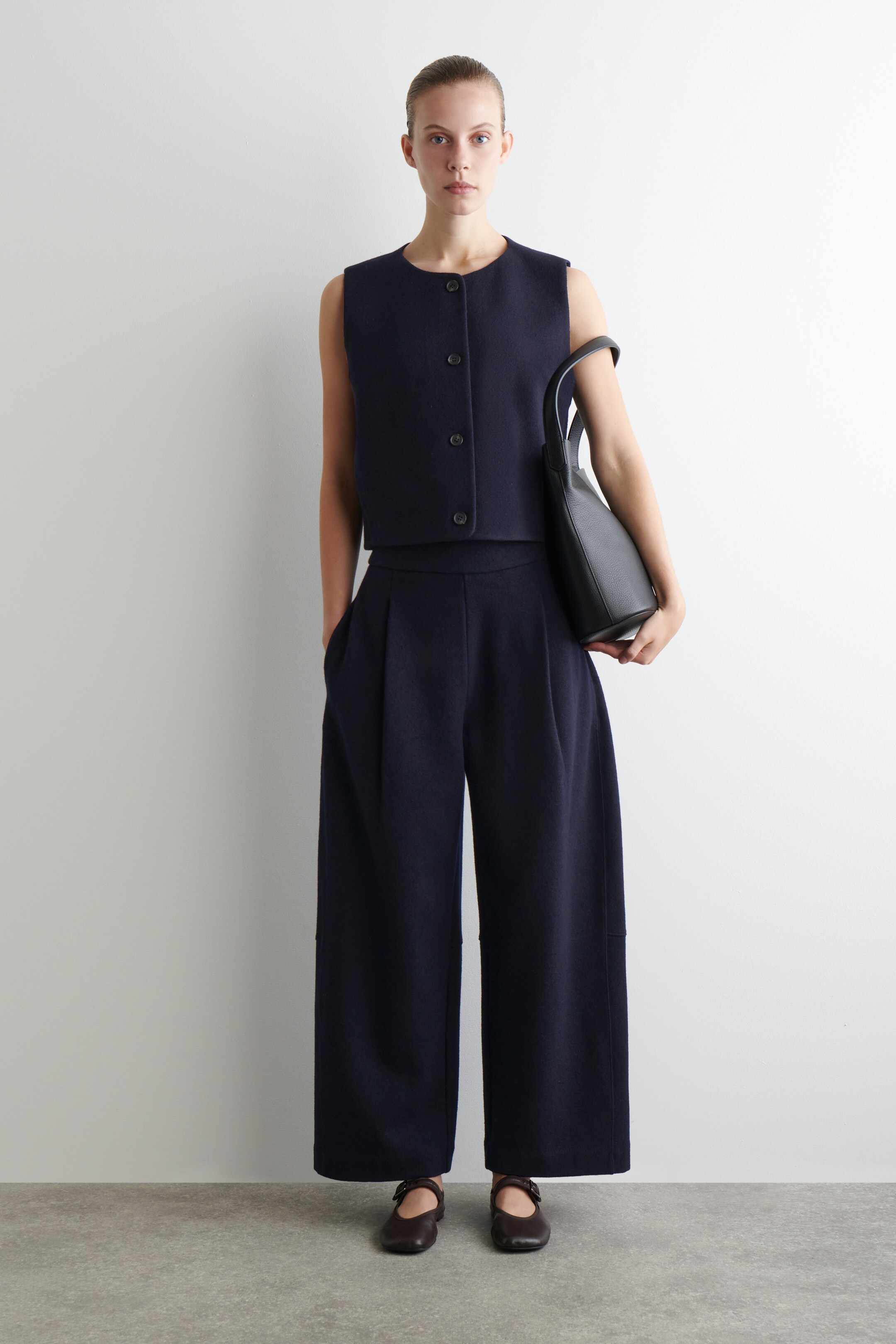 Visa större bild: En marinblå tvådelad outfit, bestående av en kortärmad väst med rund hals och tre knappar framtill, samt matchande vida byxor med veck, presenteras framifrån. En svart handväska hålls i handen.