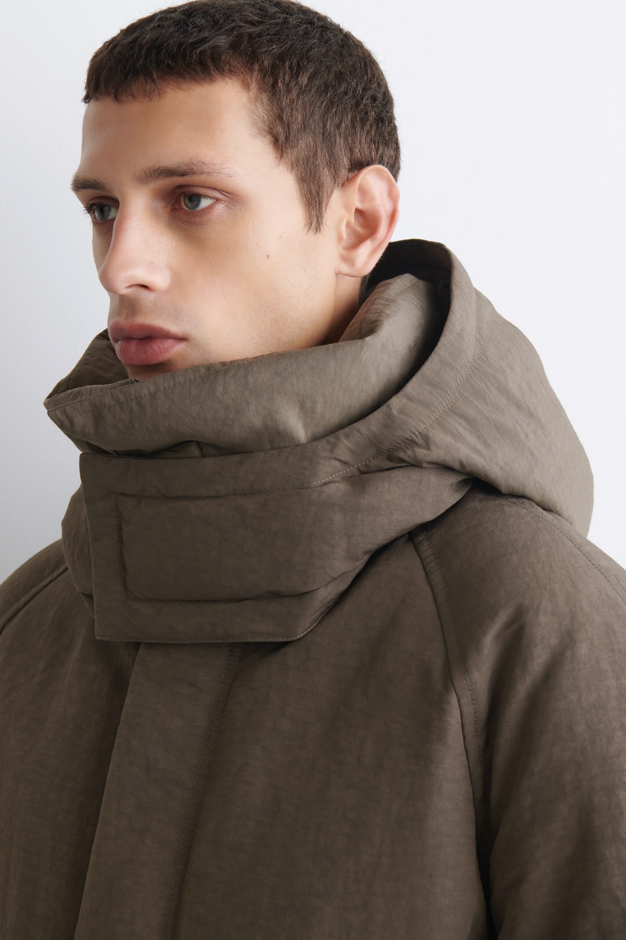 The oversized nylon-linen hooded down puffer jacket in muted brown is shown in a close-up three-quarter profile facing left, highlighting its high, padded collar and oversized hood. The jacket features a textured, matte finish with raglan shoulder seams and a concealed front closure. The oversized nylon-linen hooded down puffer jacket in muted brown is shown in a close-up three-quarter profile facing left, highlighting its high, padded collar and oversized hood. The jacket features a textured, matte finish with raglan shoulder seams and a concealed front closure.