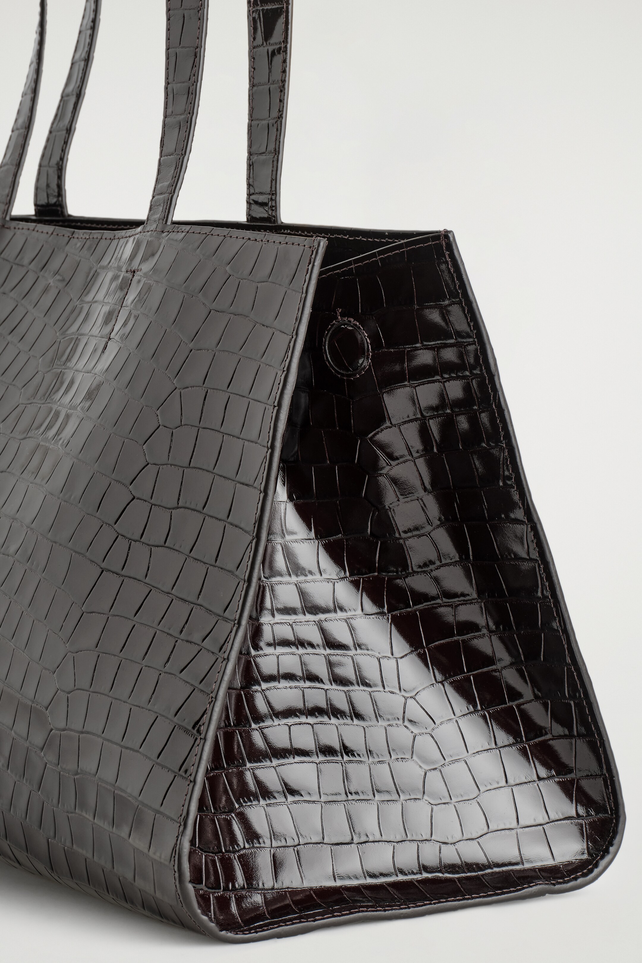 Agrandir l'image: Un sac fourre-tout en cuir gaufré croco foncé et brillant est incliné vers la gauche, avec un large panneau avant et un panneau latéral à coutures qui crée une forme cintrée, maintenu par une attache circulaire. Deux longues bandoulières s'étendent vers le haut.