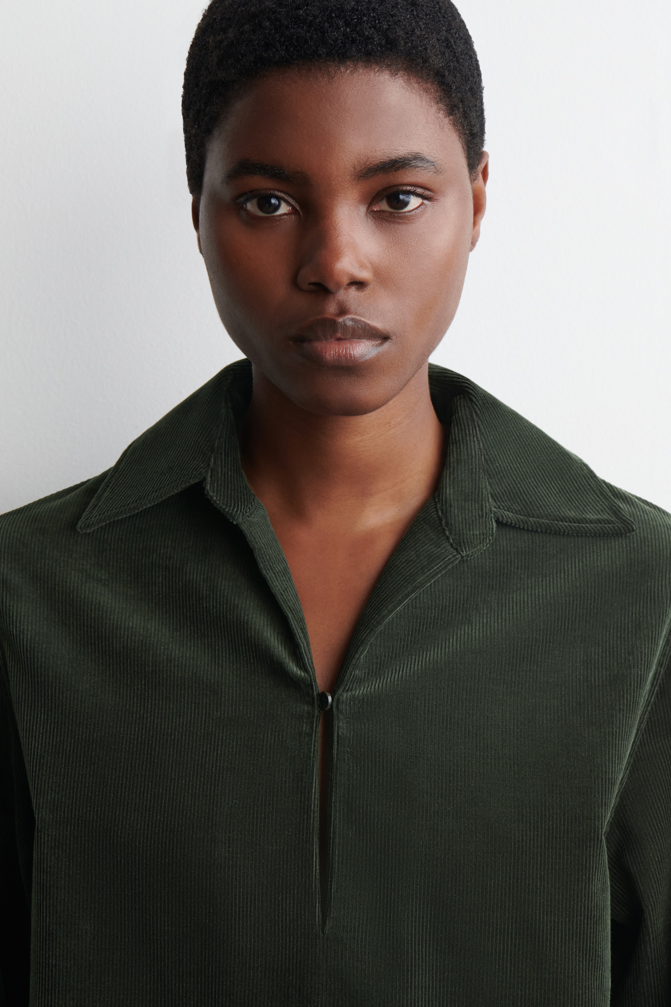 Visualizza immagine più grande: Un vestito a camicia in velluto a coste verde scuro è indossato dalla modella. Il suo colletto aperto è visibile, e la chiusura frontale è parzialmente chiusa con un bottone.