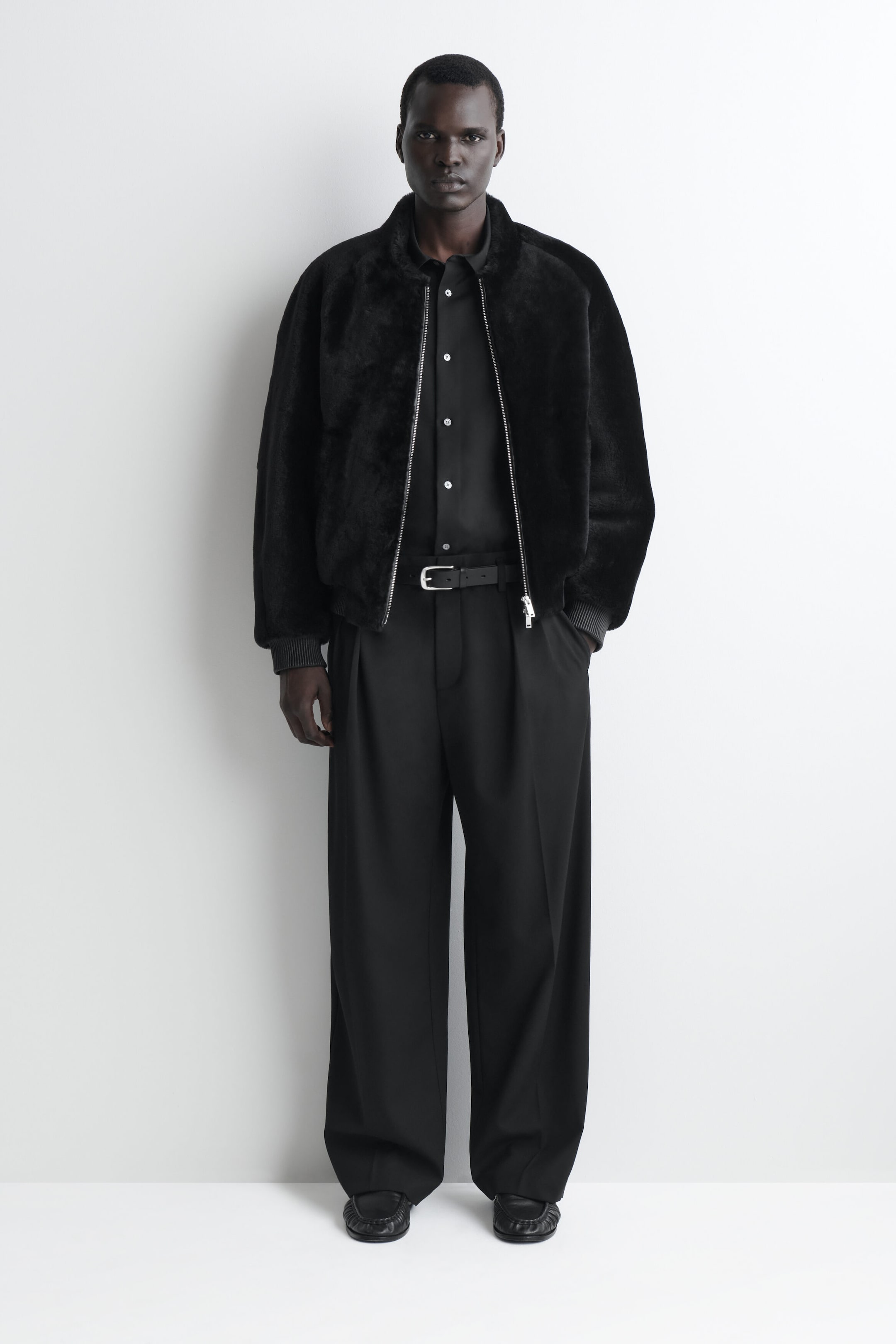 The black padded shearling bomber jacket is showcased from a front-facing angle, featuring a plush textured finish and a relaxed, voluminous silhouette. It has a high collar, ribbed knit cuffs, and a silver-tone front zipper left open over a layered black shirt.