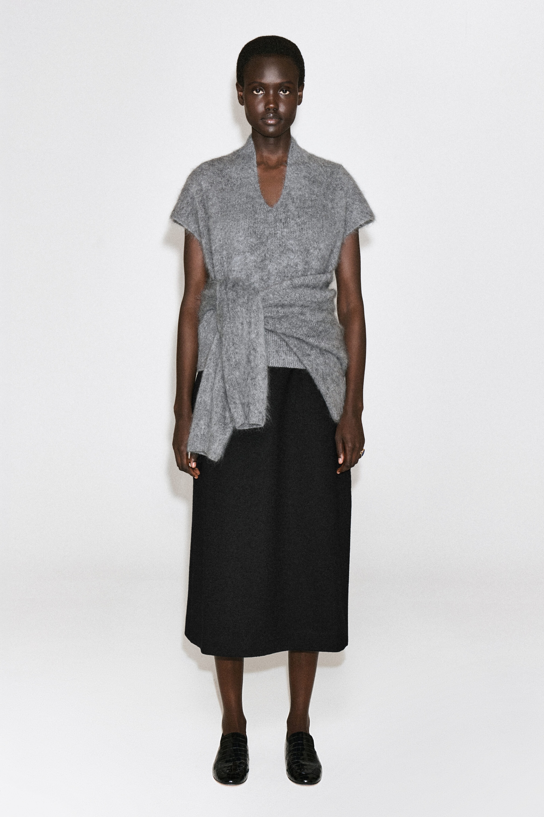 Un mannequin porte un haut gris en maille brossée, vu de face, mettant en valeur son encolure en V arrondie, ses manches courtes drapées et son détail cache-cœur à nouer à la taille. Le haut est agencé avec une jupe midi noire. Un mannequin porte un haut gris en maille brossée, vu de face, mettant en valeur son encolure en V arrondie, ses manches courtes drapées et son détail cache-cœur à nouer à la taille. Le haut est agencé avec une jupe midi noire.