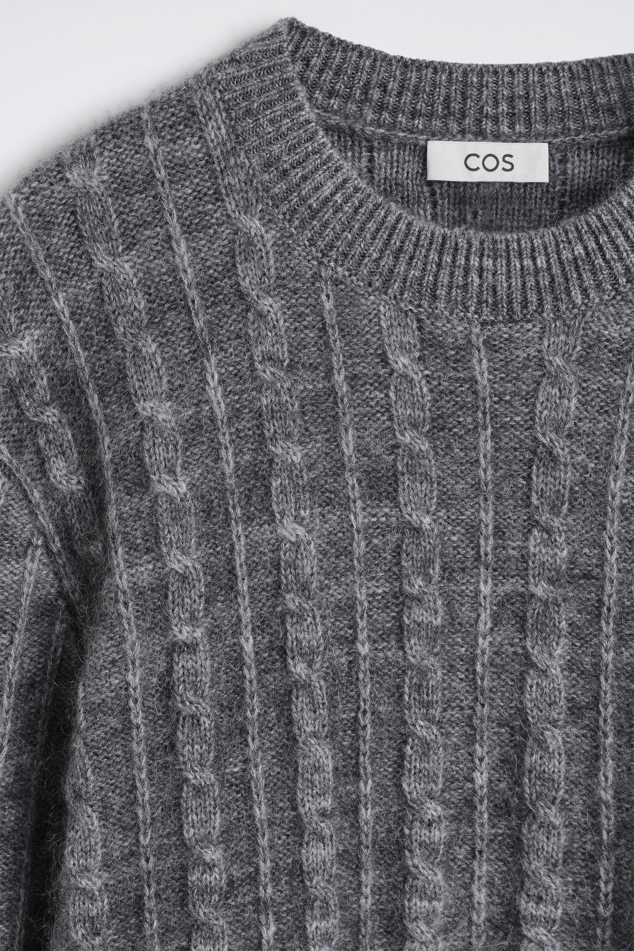 A front-facing close-up of a grey cable-knit sweater shows vertical cable patterns and a ribbed crew neck. A white label with the brand name "COS" is visible inside the collar of the textured knit garment. A front-facing close-up of a grey cable-knit sweater shows vertical cable patterns and a ribbed crew neck. A white label with the brand name "COS" is visible inside the collar of the textured knit garment.