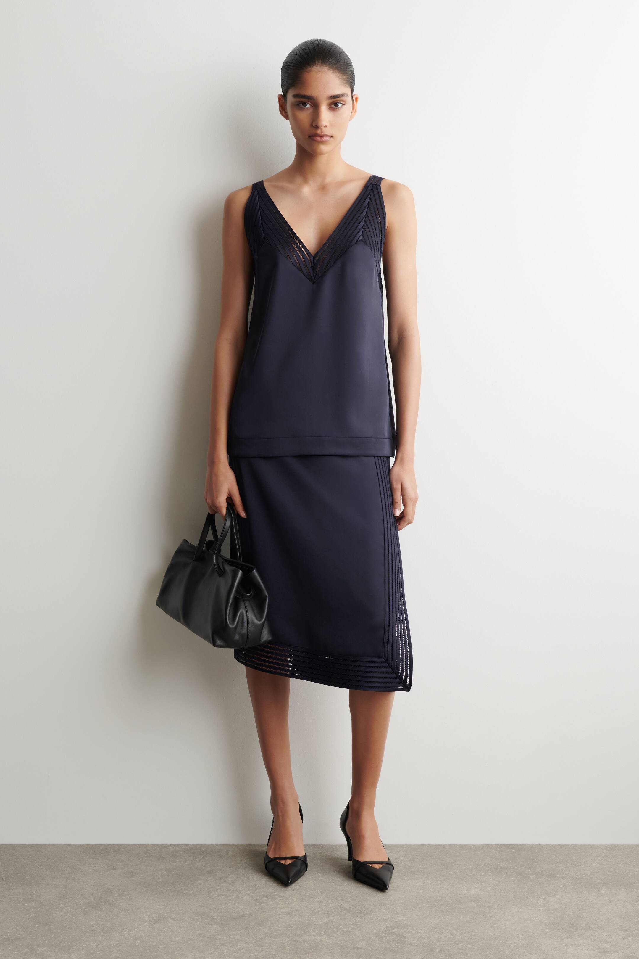 Visualizza immagine più grande: Il completo a due pezzi in blu navy presenta un top senza maniche con scollo a V e dettagli plissettati trasparenti sul petto. La gonna midi coordinata è progettata con pannelli a righe trasparenti orizzontali e verticali vicino all'orlo.