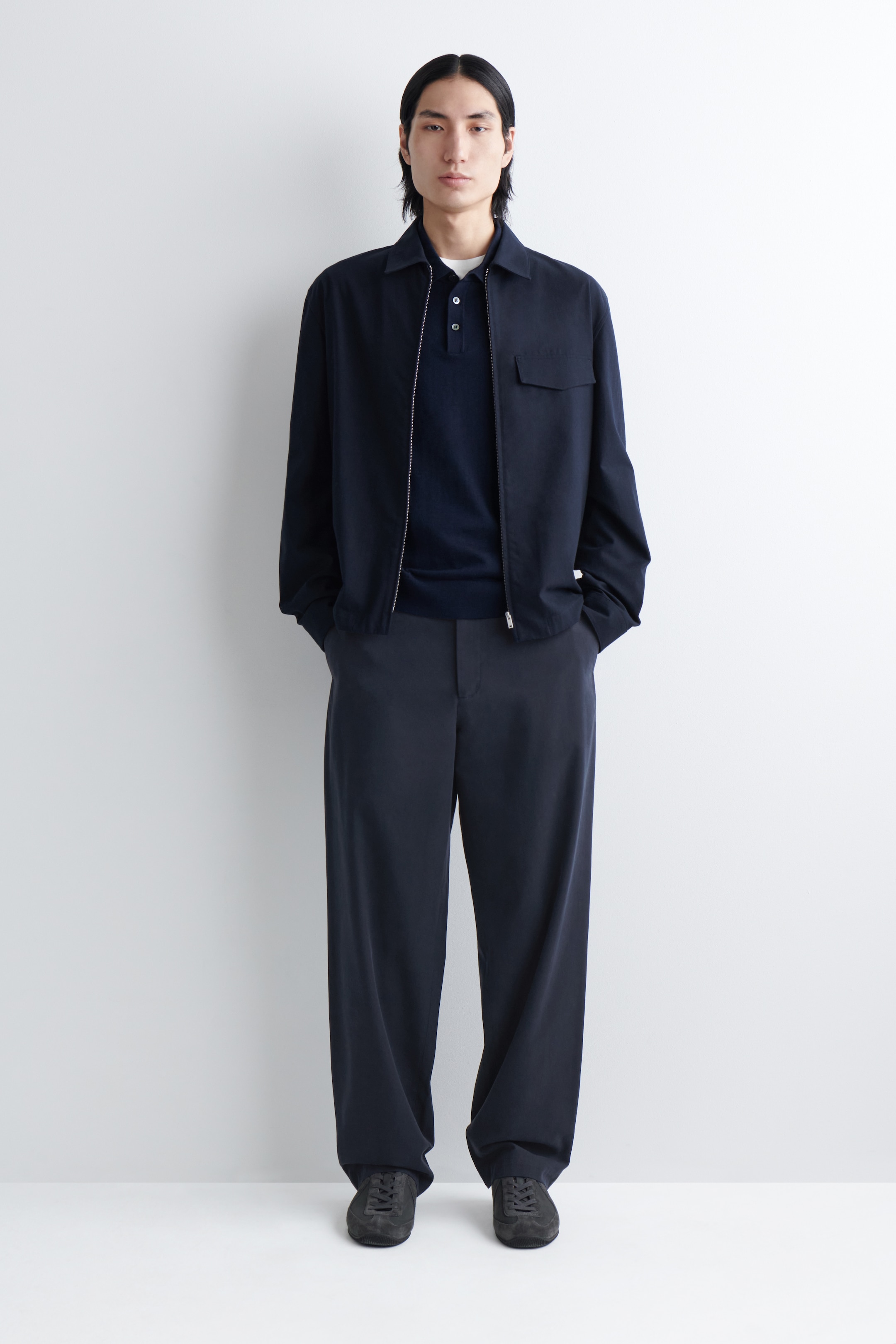 Grotere afbeelding bekijken: Een marineblauwe jas met ritssluiting wordt gedragen, voorzien van een klassieke kraag en een enkele klepzak op de linkerborst. Deze is gelaagd over een marineblauw poloshirt met een drieknoopsluiting en wordt gecombineerd met wijde marineblauwe broek en donkere vetersneakers, allemaal gepresenteerd in een vooraanzicht.