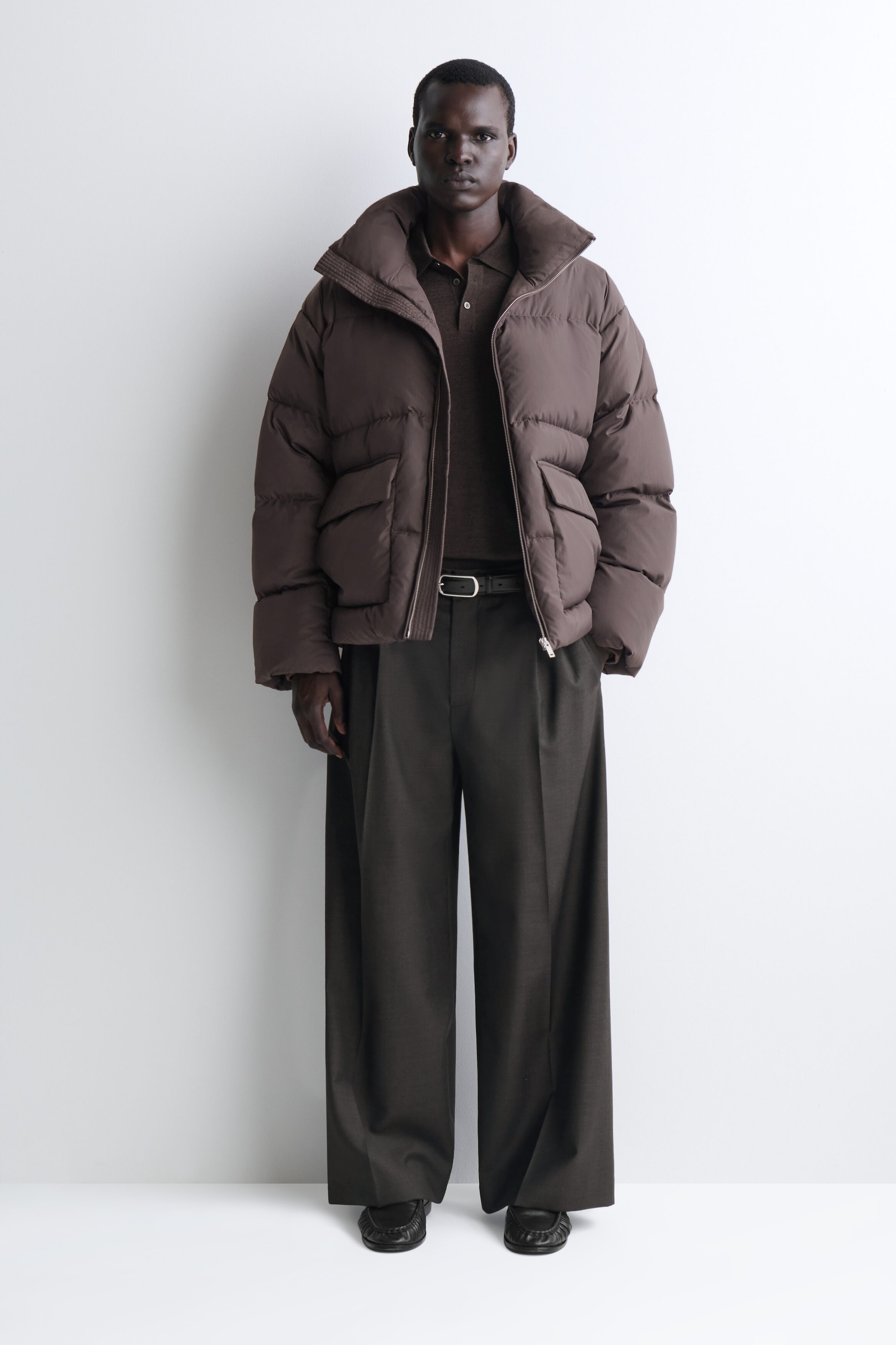 A model is wearing a dark brown funnel-neck nylon down puffer jacket in a front-facing view, showcasing its relaxed, horizontally quilted design. The jacket features a high, structured funnel collar, an open front zipper closure, and two large flap pockets at the waist. A model is wearing a dark brown funnel-neck nylon down puffer jacket in a front-facing view, showcasing its relaxed, horizontally quilted design. The jacket features a high, structured funnel collar, an open front zipper closure, and two large flap pockets at the waist.