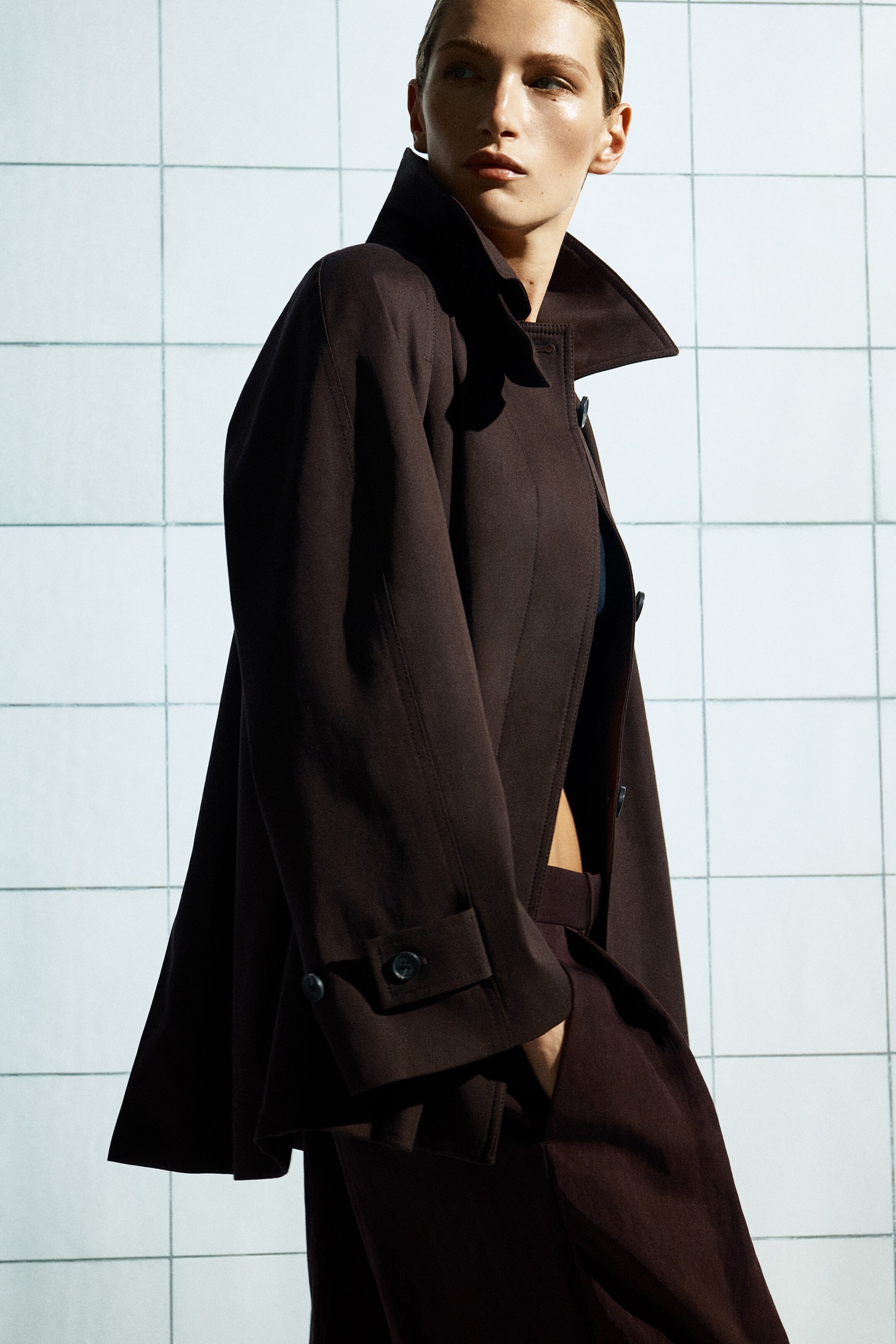 Größeres Bild anzeigen: Ein dunkelbrauner A-Linien-Trenchcoat wird getragen, wobei der spitze Kragen aufgestellt ist und eine verdeckte Knopfleiste sowie geknöpfte Manschettenriemen aufweist. Er ist über passenden dunkelbraunen Hosen mit weitem Bein und sichtbaren Vorderfalten geschichtet. Der Mantel ist teilweise offen und gibt den Blick auf die Hosen frei.