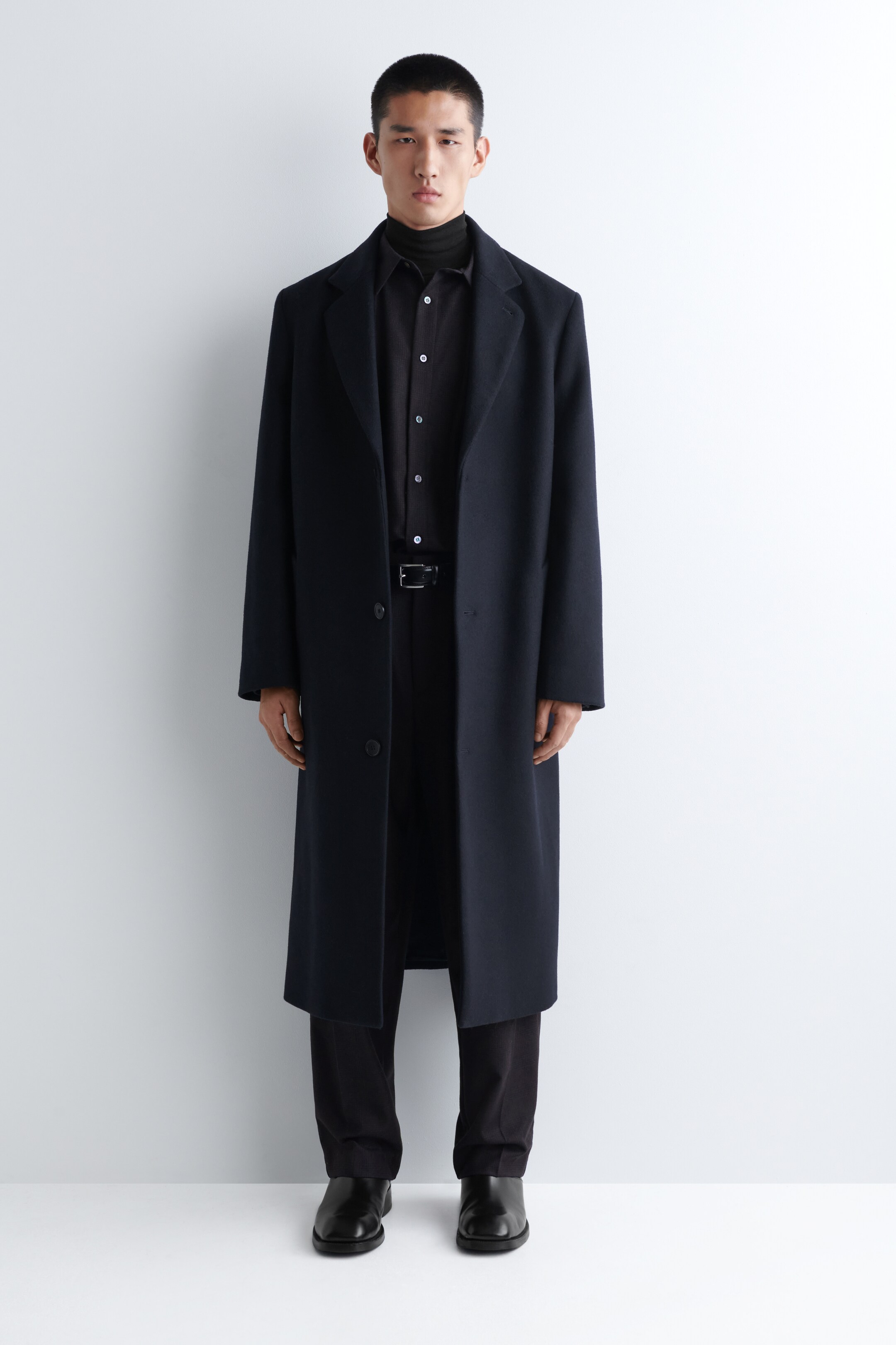Agrandir l'image: Un long manteau bleu marine à simple boutonnage est orienté vers l'avant, avec des revers crantés et deux boutons avant visibles. Il présente une coupe décontractée et est porté ouvert sur une chemise noire à col et un col roulé.