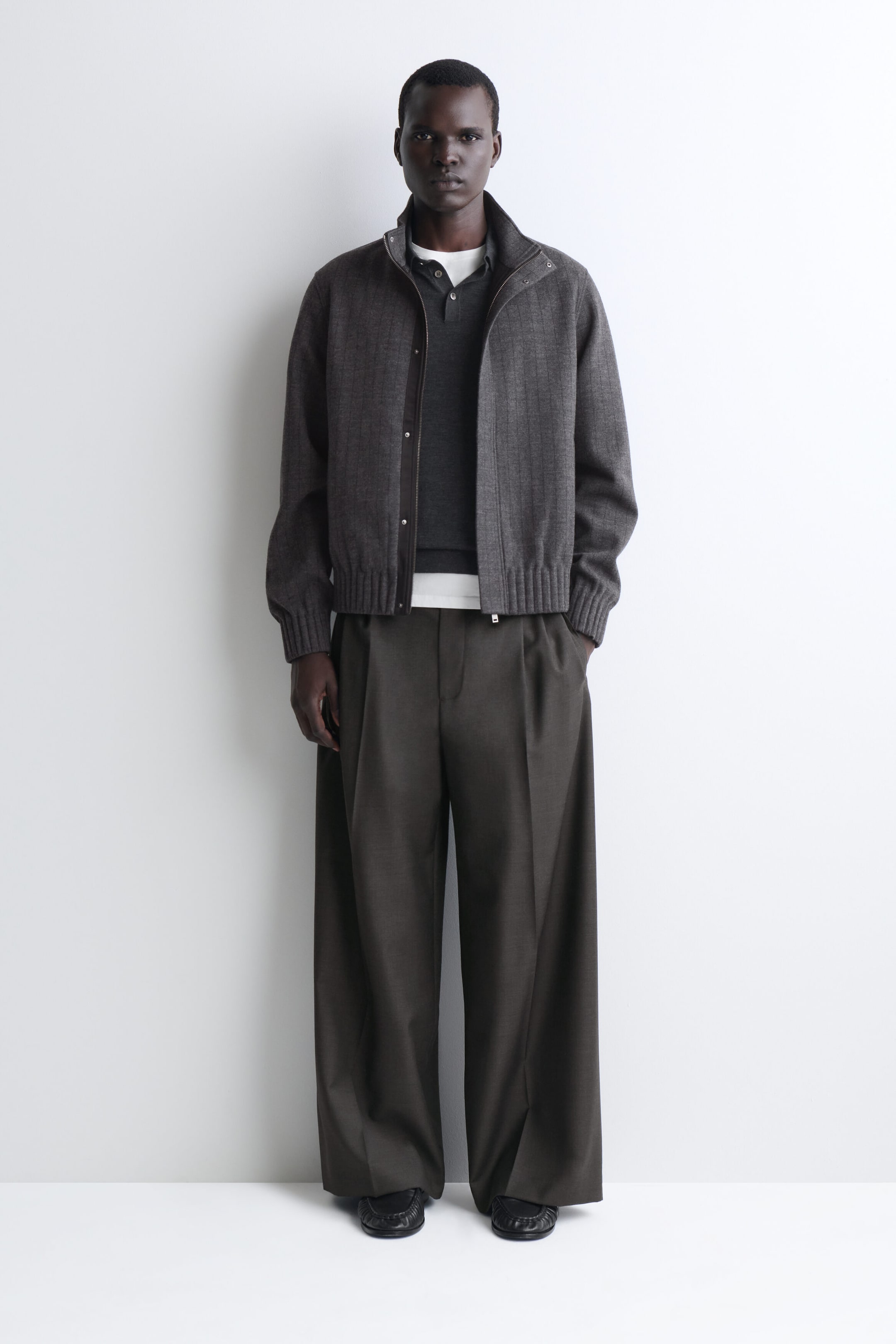 A model is wearing a front-facing outfit featuring a textured gray zip-up jacket with a high collar, ribbed cuffs, and a ribbed hem. The jacket is layered over a dark gray polo shirt and a white T-shirt, paired with dark brown, wide-leg pleated trousers. A model is wearing a front-facing outfit featuring a textured gray zip-up jacket with a high collar, ribbed cuffs, and a ribbed hem. The jacket is layered over a dark gray polo shirt and a white T-shirt, paired with dark brown, wide-leg pleated trousers.