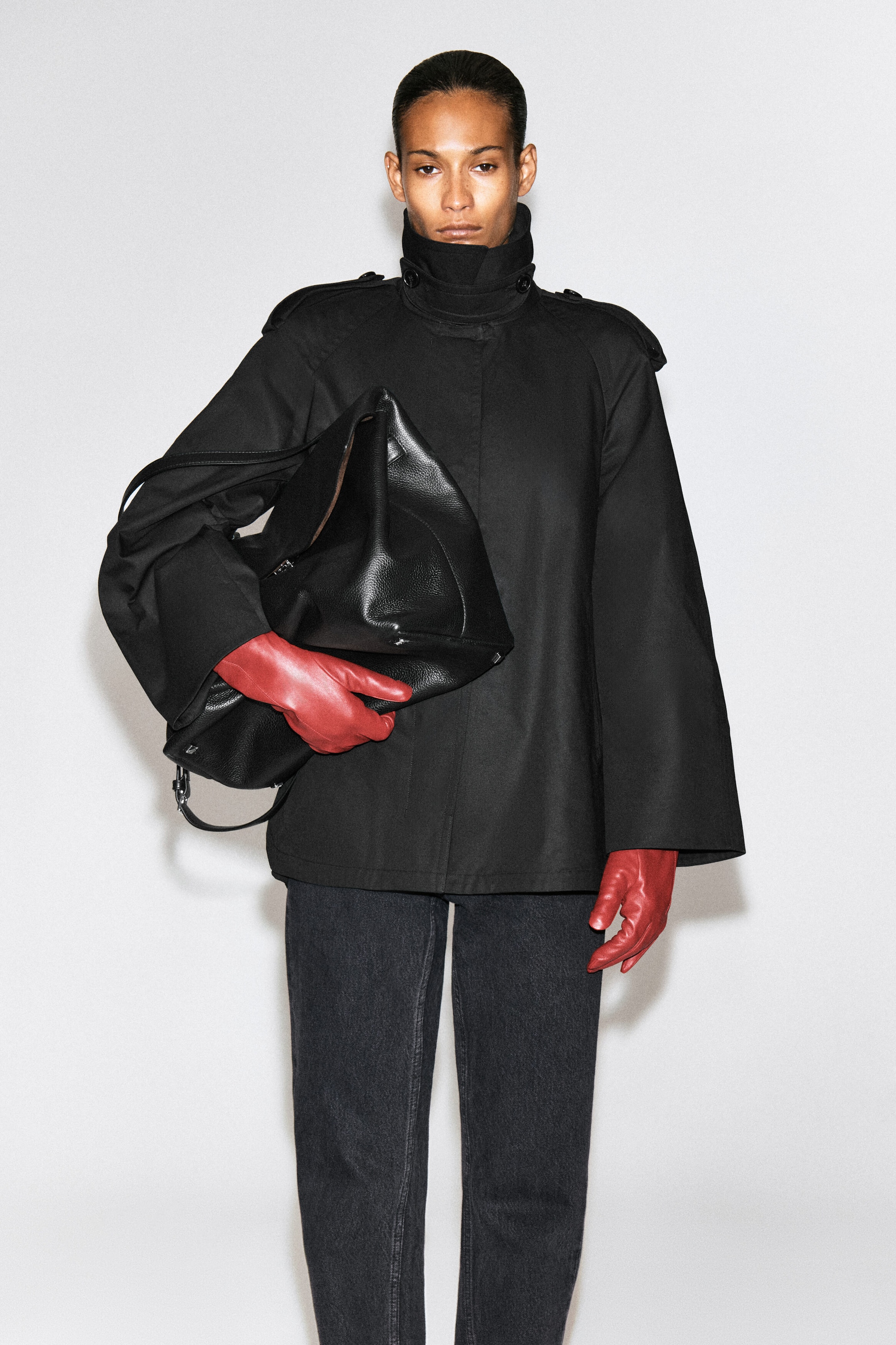 La mannequin porte une veste noire à col montant et à manches larges, agencée à un jean gris foncé et à des gants en cuir rouge. Un grand sac en cuir noir texturé est tenu sous le bras de la mannequin. La mannequin porte une veste noire à col montant et à manches larges, agencée à un jean gris foncé et à des gants en cuir rouge. Un grand sac en cuir noir texturé est tenu sous le bras de la mannequin.