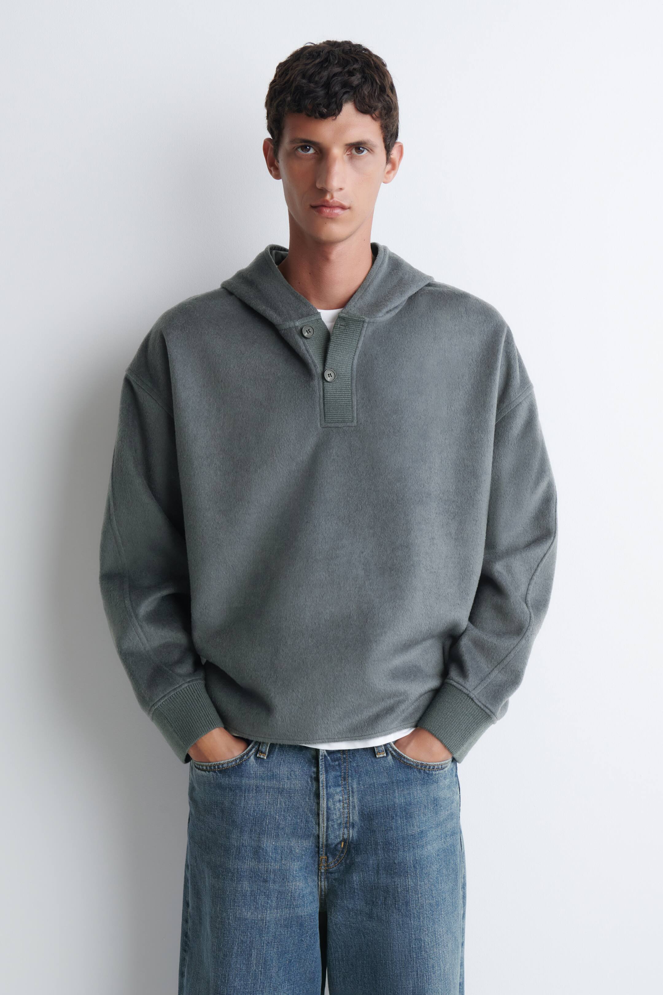 Agrandir l'image: Un pull à capuche vert grisâtre avec encolure henley boutonnée et poignets côtelés est porté par le mannequin. Le haut à coupe décontractée est superposé à un t-shirt blanc et associé à un jean bleu.