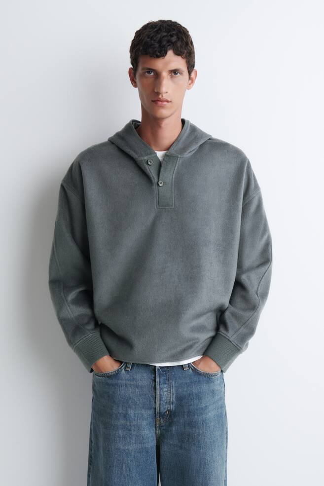 Ein graugrÃ¼nlicher Kapuzenpullover mit geknÃ¶pftem Henley-Ausschnitt und gerippten BÃ¼ndchen wird vom Model getragen. Das Oberteil in entspannter Passform ist Ã¼ber einem weiÃen T-Shirt getragen und mit blauen Jeans kombiniert. - 1