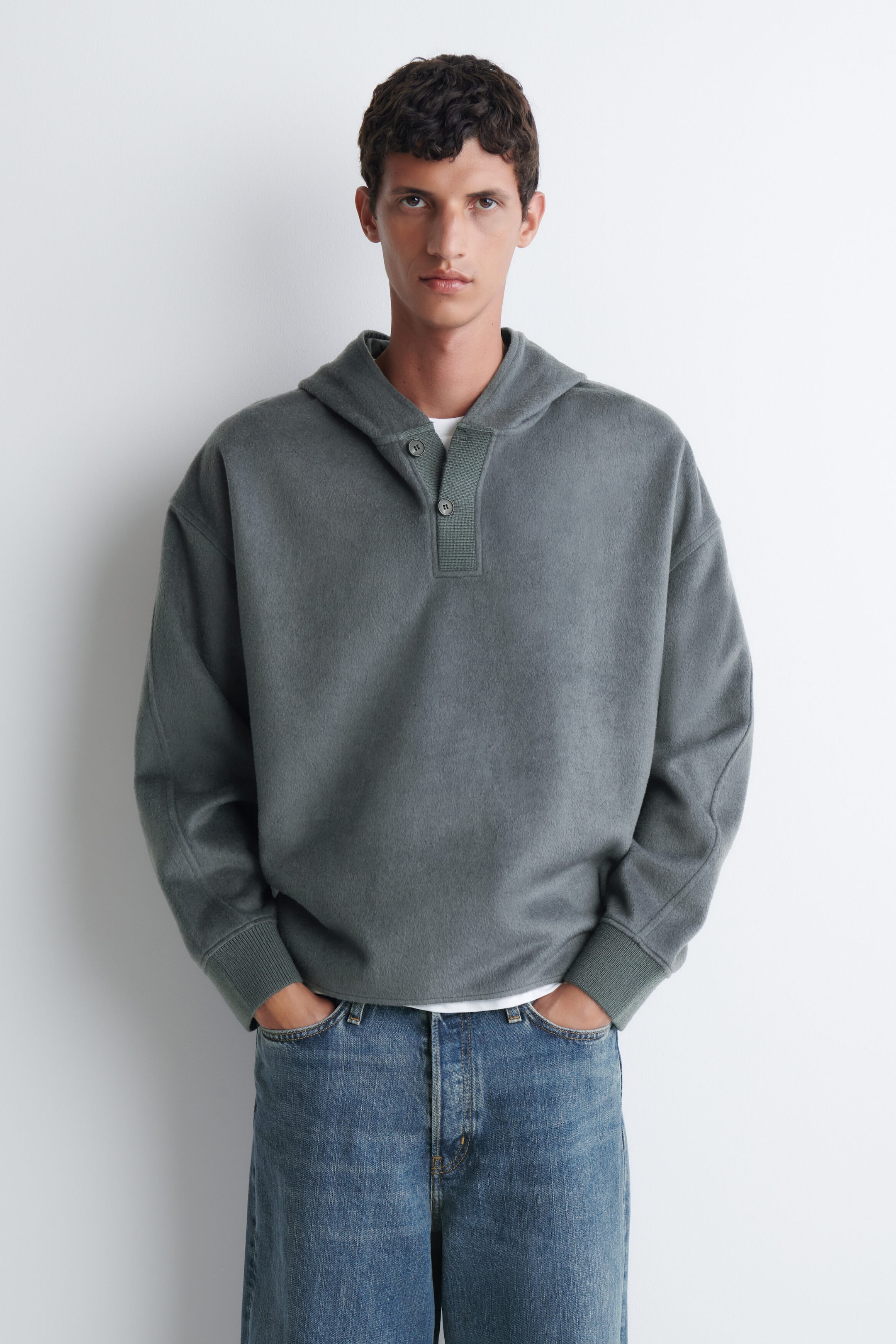 Un pull à capuche vert grisâtre avec encolure henley boutonnée et poignets côtelés est porté par le mannequin. Le haut à coupe décontractée est superposé à un t-shirt blanc et assorti à un jean bleu. Un pull à capuche vert grisâtre avec encolure henley boutonnée et poignets côtelés est porté par le mannequin. Le haut à coupe décontractée est superposé à un t-shirt blanc et assorti à un jean bleu.