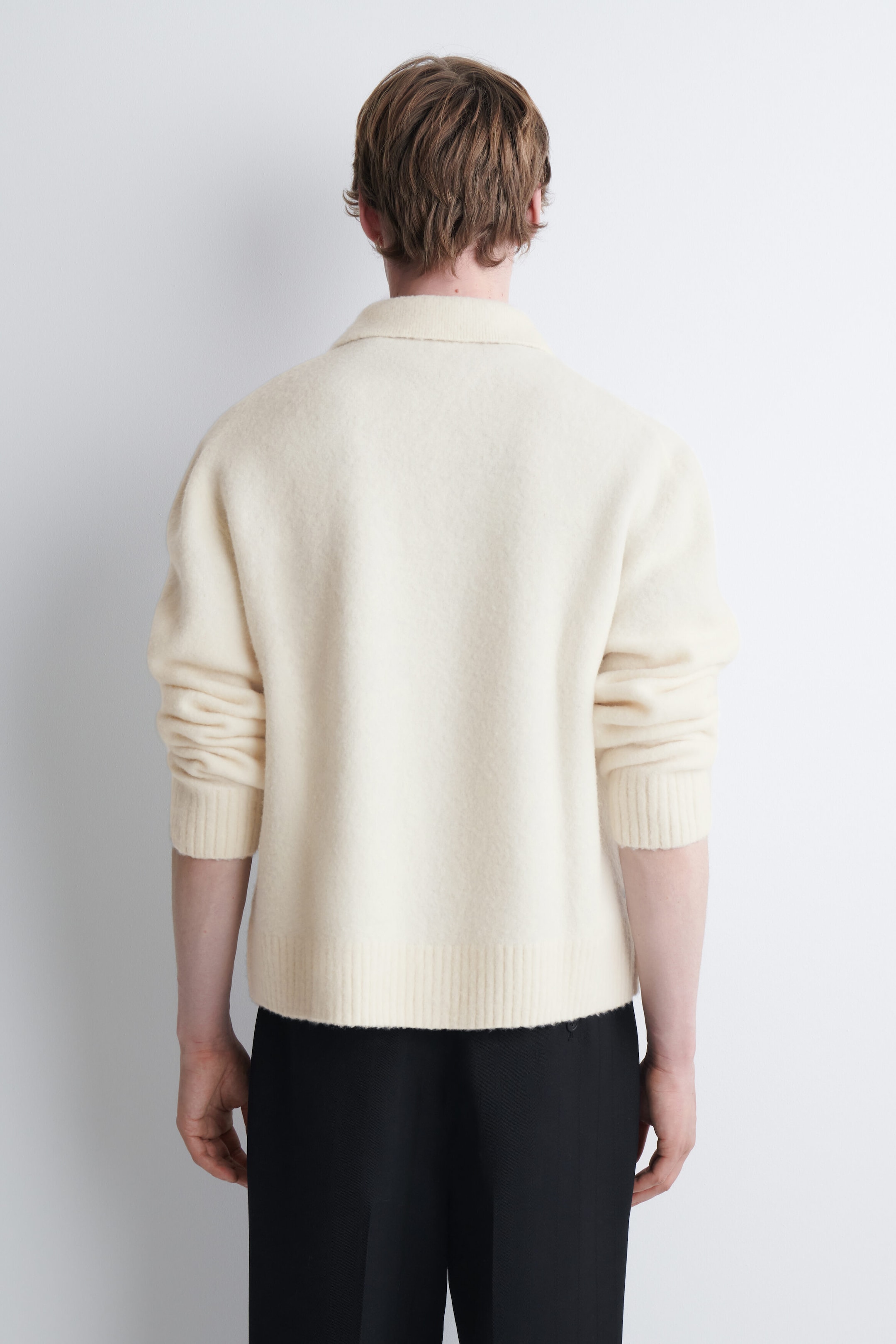 A brushed wool polo shirt in an off-white hue is worn by the model, viewed from the back. The long-sleeved shirt features a soft, textured finish, a polo collar, and ribbed cuffs and hem, and is styled with black trousers.