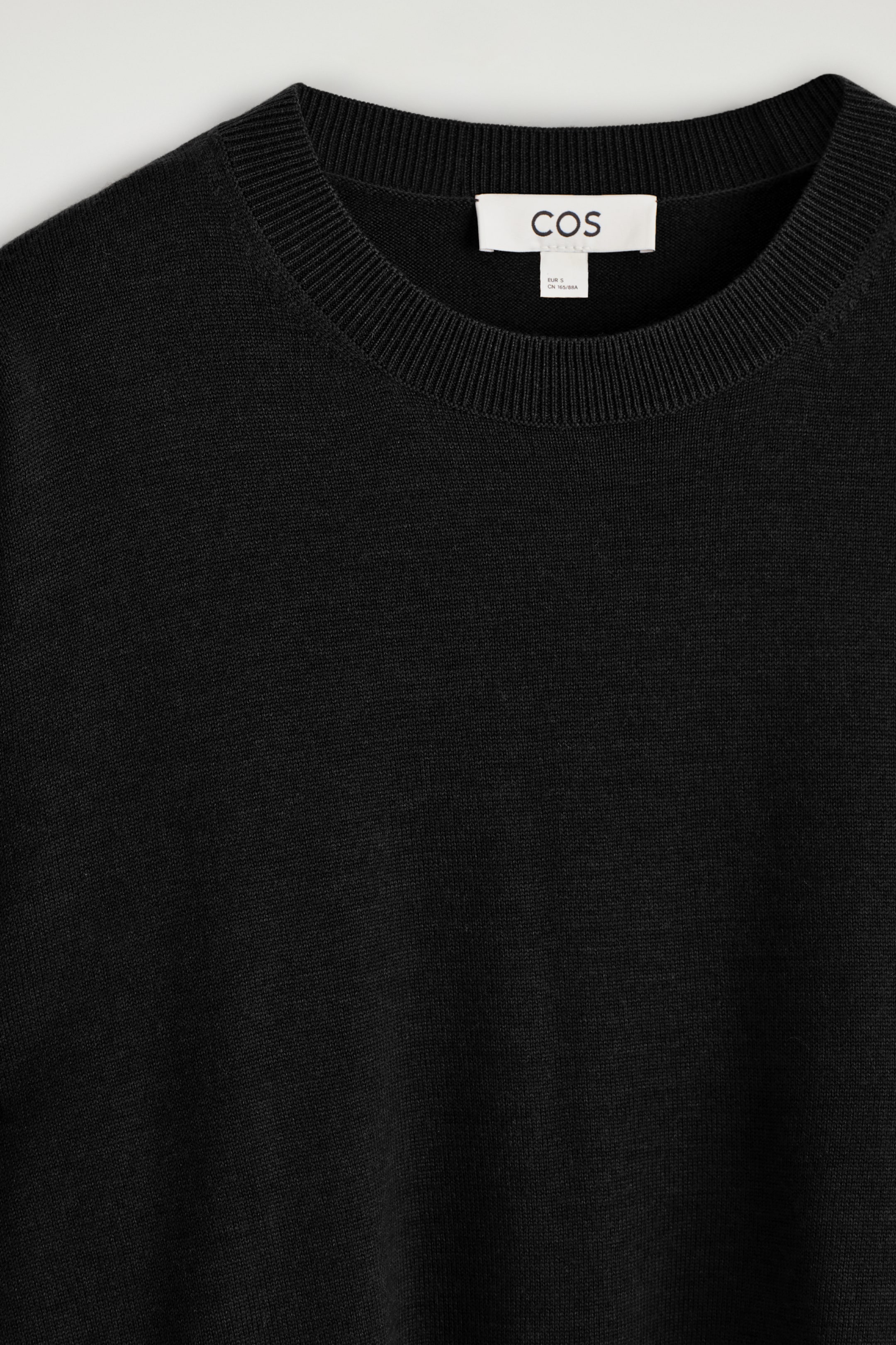 Ver imagen más grande: Un jersey de punto negro se muestra en una vista frontal de cerca, mostrando su cuello redondo acanalado y su textura finamente tejida. Una etiqueta blanca de la marca COS es visible dentro del escote.