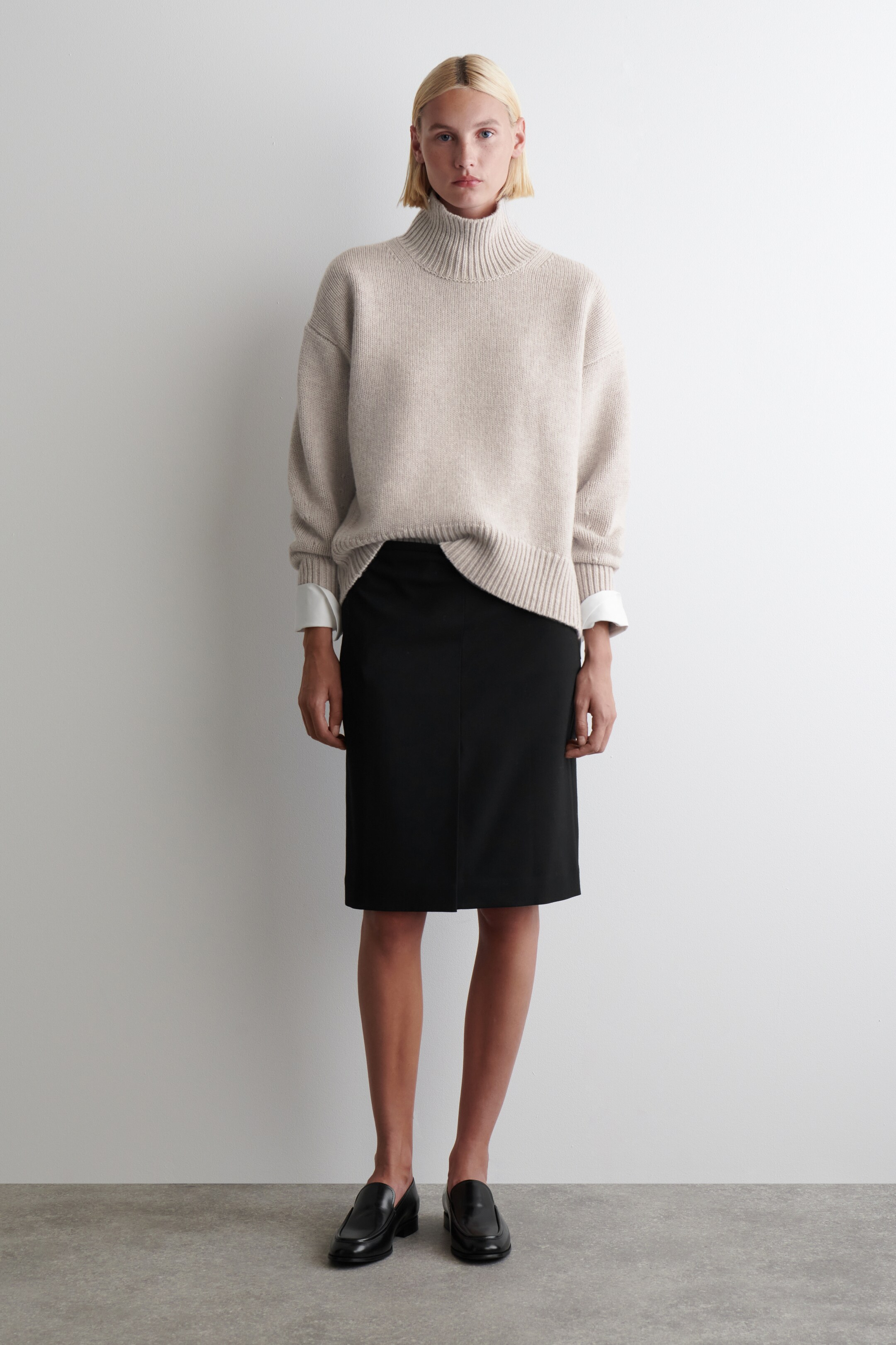 Grösseres Bild ansehen: Ein beige gerippter Rollkragenpullover zeichnet sich durch eine lockere Passform mit gerippten Bündchen und Saum aus und wird über einem weißen Hemd getragen, dessen Manschetten sichtbar sind. Er wird mit einem schwarzen knielangen Bleistiftrock und schwarzen Slipper-Loafern kombiniert.