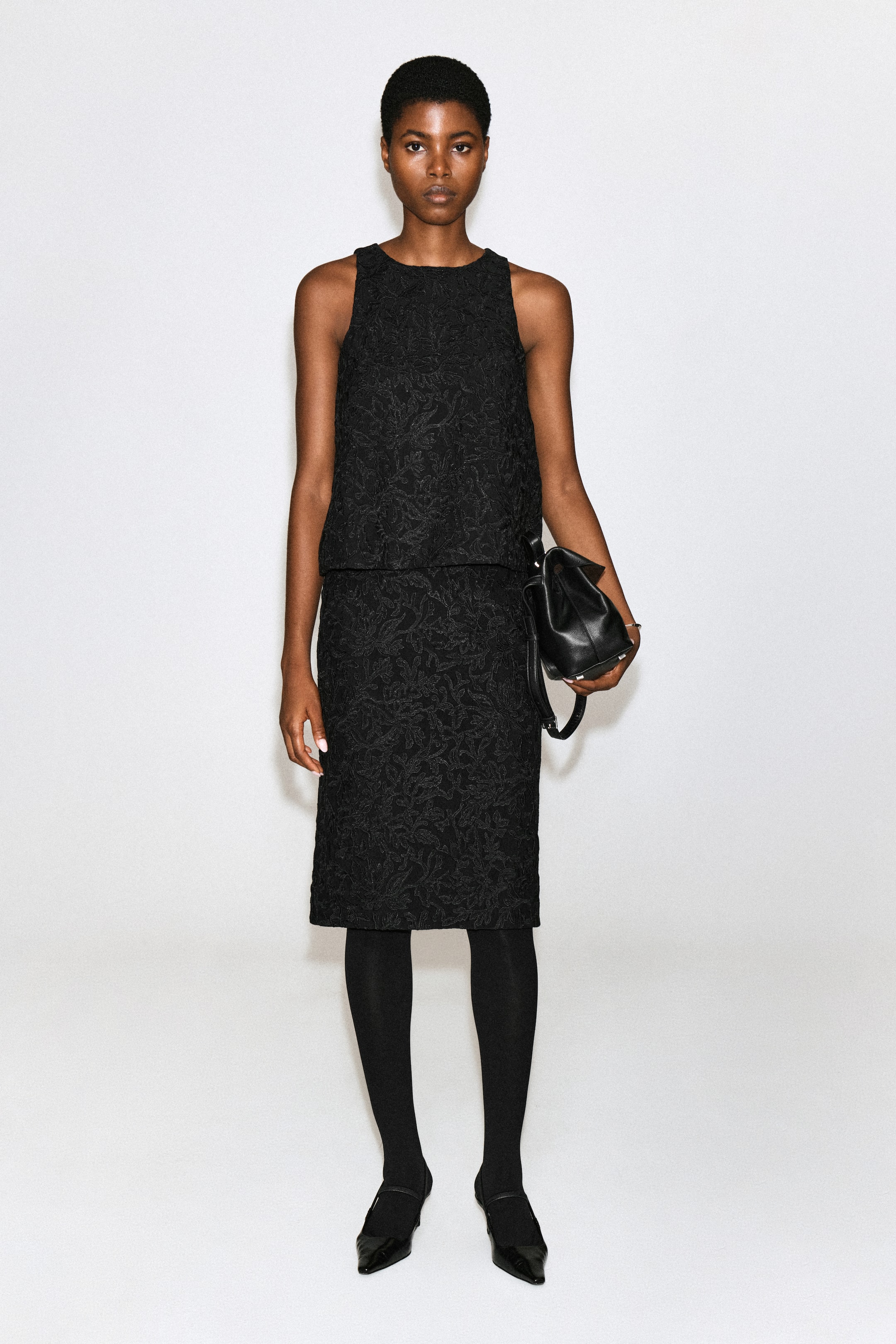 View larger image: A black midi skirt featuring textured floral lace embroidery is being worn by the model. The skirt is styled with a matching sleeveless top, black tights, and black pointed-toe flat shoes, while a black leather handbag is being held by the model.