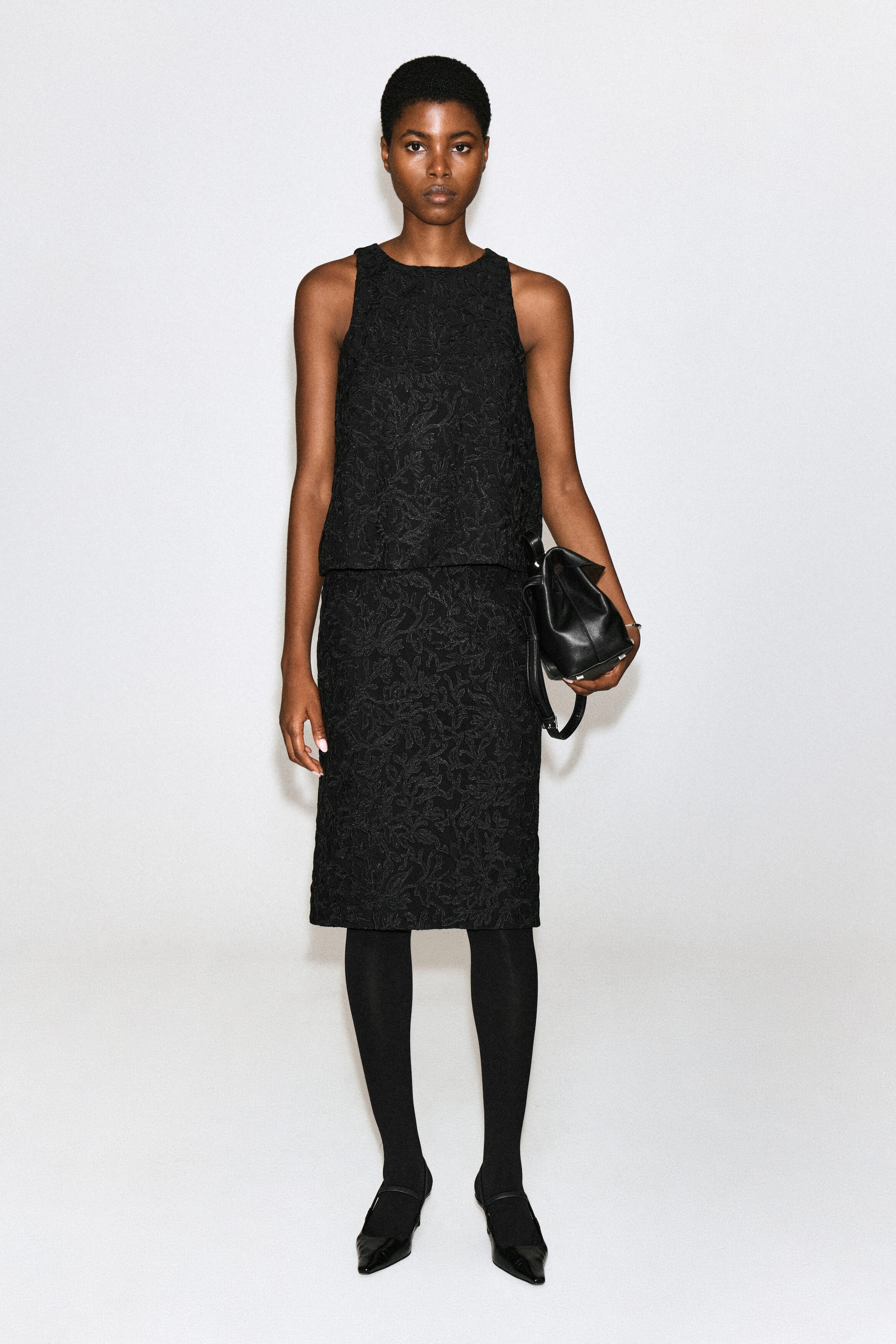 A black midi skirt featuring textured floral lace embroidery is being worn by the model. The skirt is styled with a matching sleeveless top, black tights, and black pointed-toe flat shoes, while a black leather handbag is being held by the model. A black midi skirt featuring textured floral lace embroidery is being worn by the model. The skirt is styled with a matching sleeveless top, black tights, and black pointed-toe flat shoes, while a black leather handbag is being held by the model.