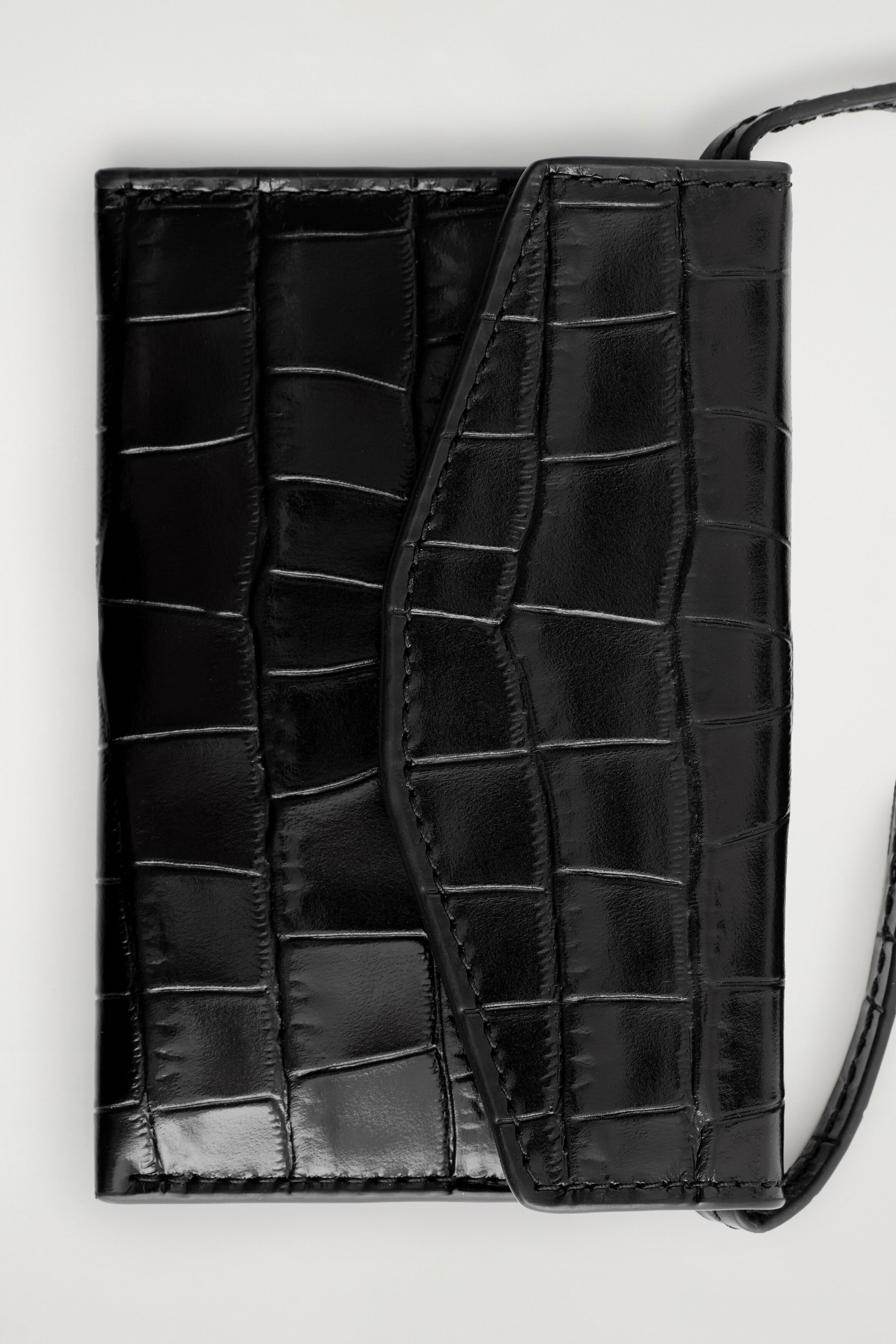 A black leather card holder is lying flat in a front-facing view, showcasing a glossy crocodile-embossed texture and a curved fold-over flap with tonal stitching. A thin matching leather strap extends from the side. A black leather card holder is lying flat in a front-facing view, showcasing a glossy crocodile-embossed texture and a curved fold-over flap with tonal stitching. A thin matching leather strap extends from the side.