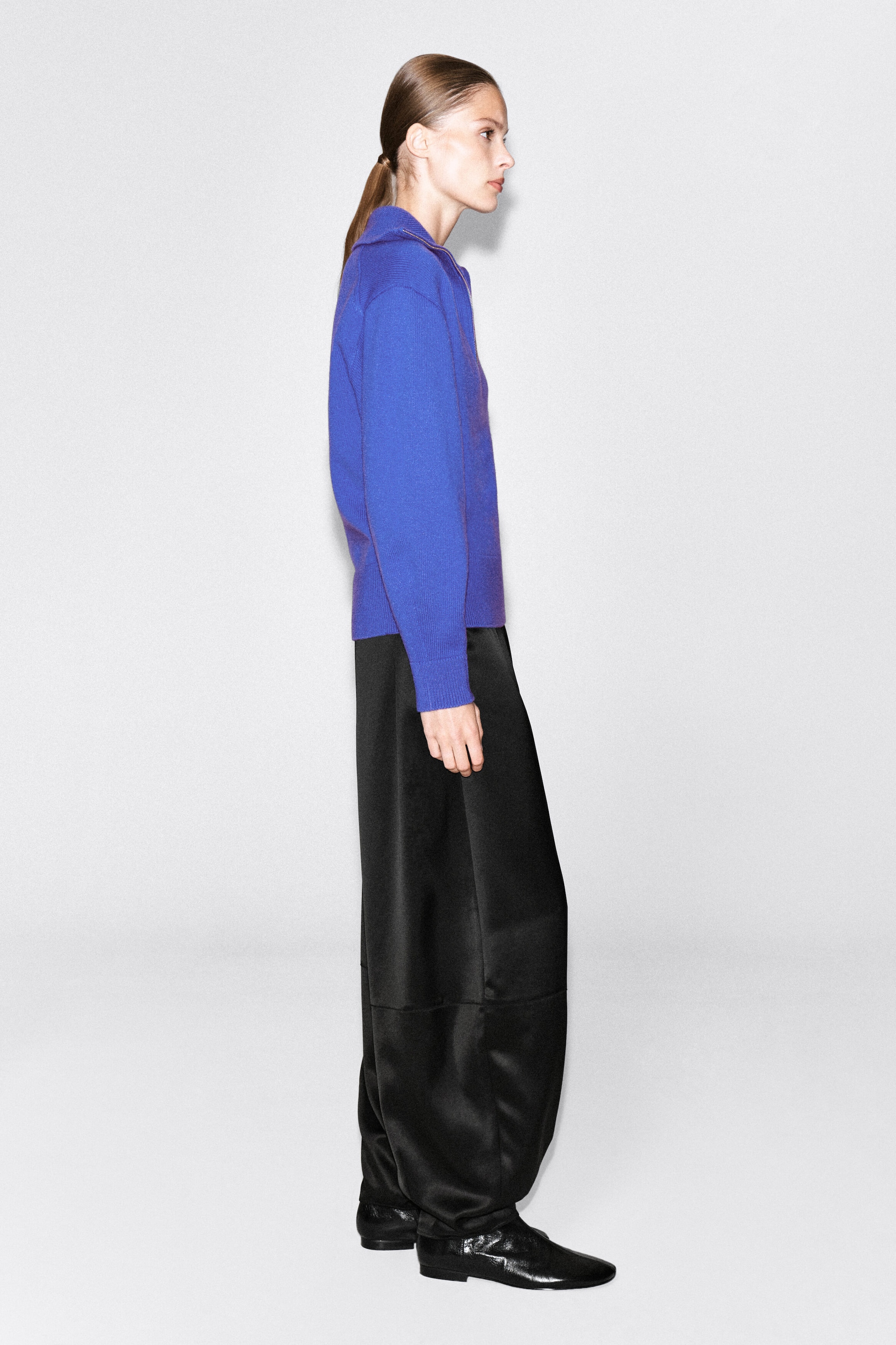 A bright blue knitted sweater is being worn by the model, featuring a funnel neckline and relaxed sleeves. The sweater is paired with black wide-leg trousers and black shoes, shown from a side-profile view. A bright blue knitted sweater is being worn by the model, featuring a funnel neckline and relaxed sleeves. The sweater is paired with black wide-leg trousers and black shoes, shown from a side-profile view.