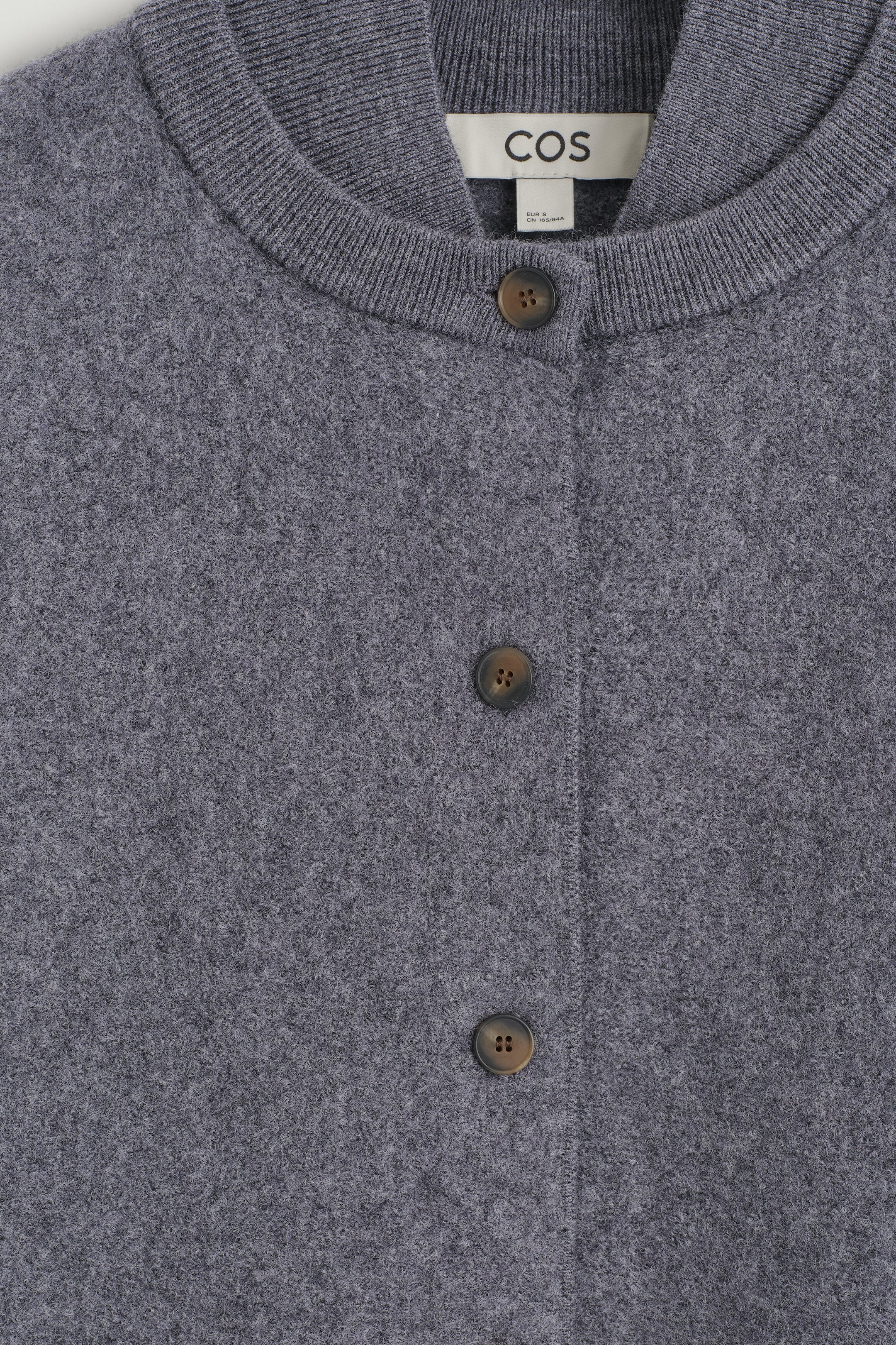 A gray boiled wool cardigan with a layered-effect ribbed collar and dark brown buttons shown in a close-up front view, highlighting the dense texture of the knit fabric. A gray boiled wool cardigan with a layered-effect ribbed collar and dark brown buttons shown in a close-up front view, highlighting the dense texture of the knit fabric.