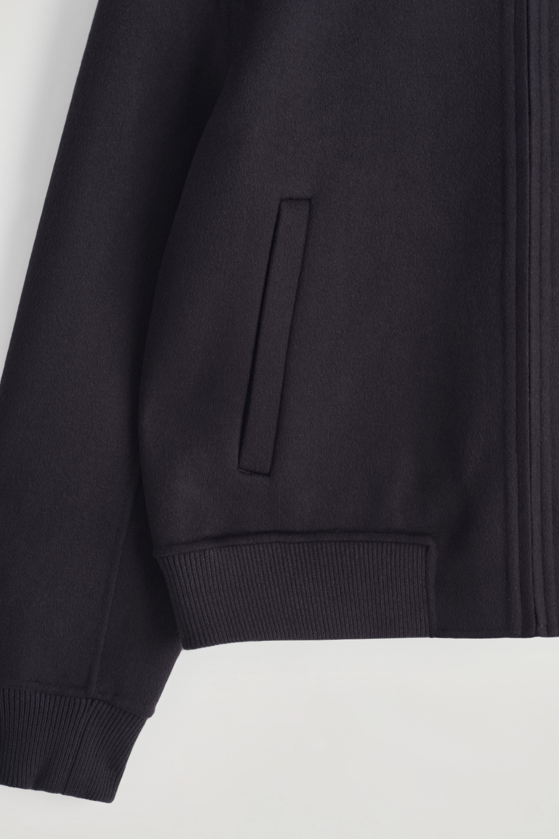 View larger image: A dark-colored bomber jacket is being shown from the front, featuring a smooth main fabric, a vertical slit pocket, ribbed cuffs, and a ribbed hem, with vertical stitching visible along the right side of the front.