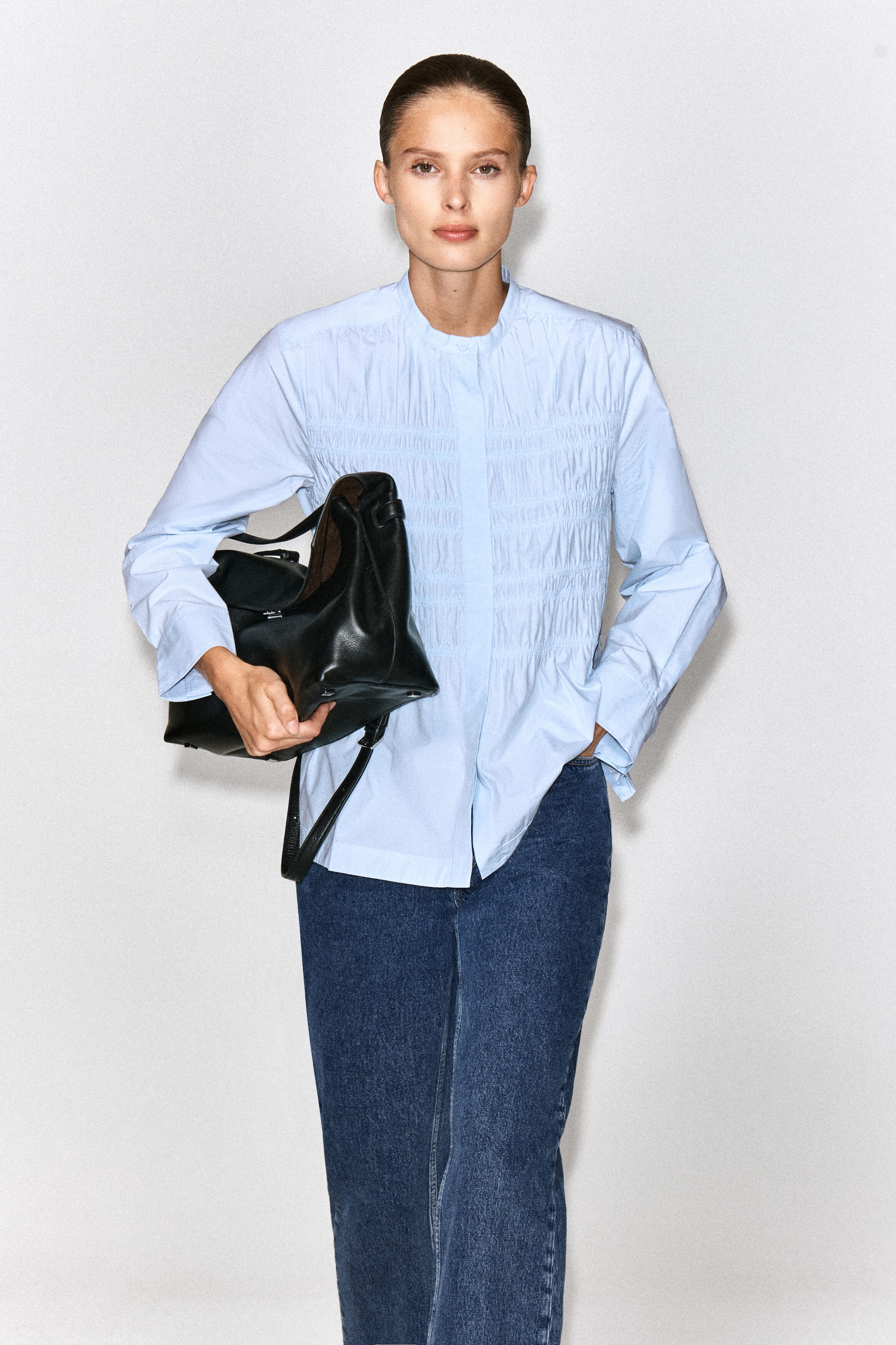 A light blue cotton blouse featuring a band collar and vertical ruched detailing across the chest is being worn by the model. The blouse is styled with blue jeans, with a black leather handbag being carried under the model's arm. A light blue cotton blouse featuring a band collar and vertical ruched detailing across the chest is being worn by the model. The blouse is styled with blue jeans, with a black leather handbag being carried under the model's arm.