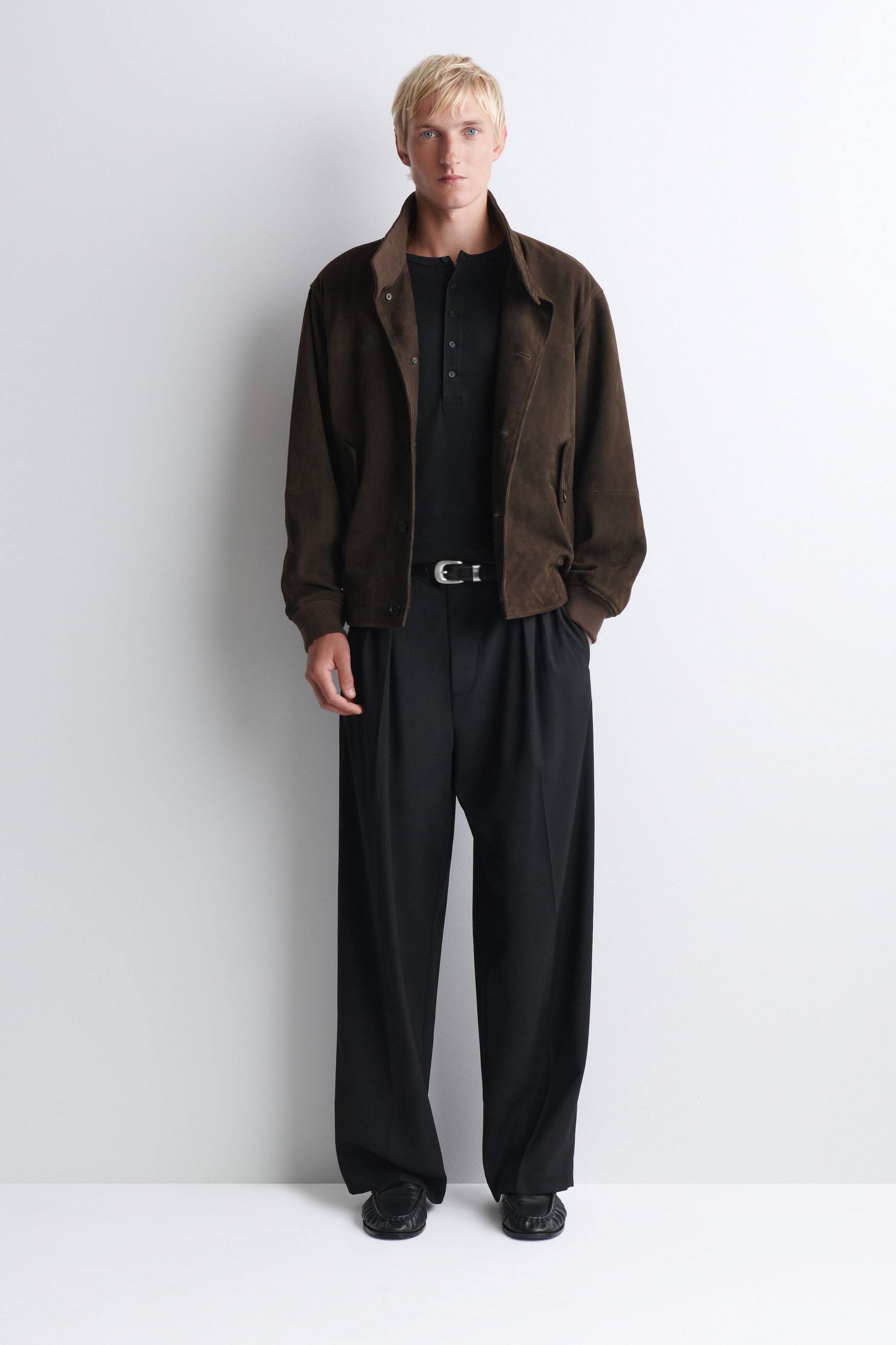 The slim ribbed cotton long-sleeved Henley T-shirt in black is facing forward, featuring a buttoned placket at the neckline. The slim-fitting shirt is worn tucked into black trousers and layered under an open brown jacket. The slim ribbed cotton long-sleeved Henley T-shirt in black is facing forward, featuring a buttoned placket at the neckline. The slim-fitting shirt is worn tucked into black trousers and layered under an open brown jacket.