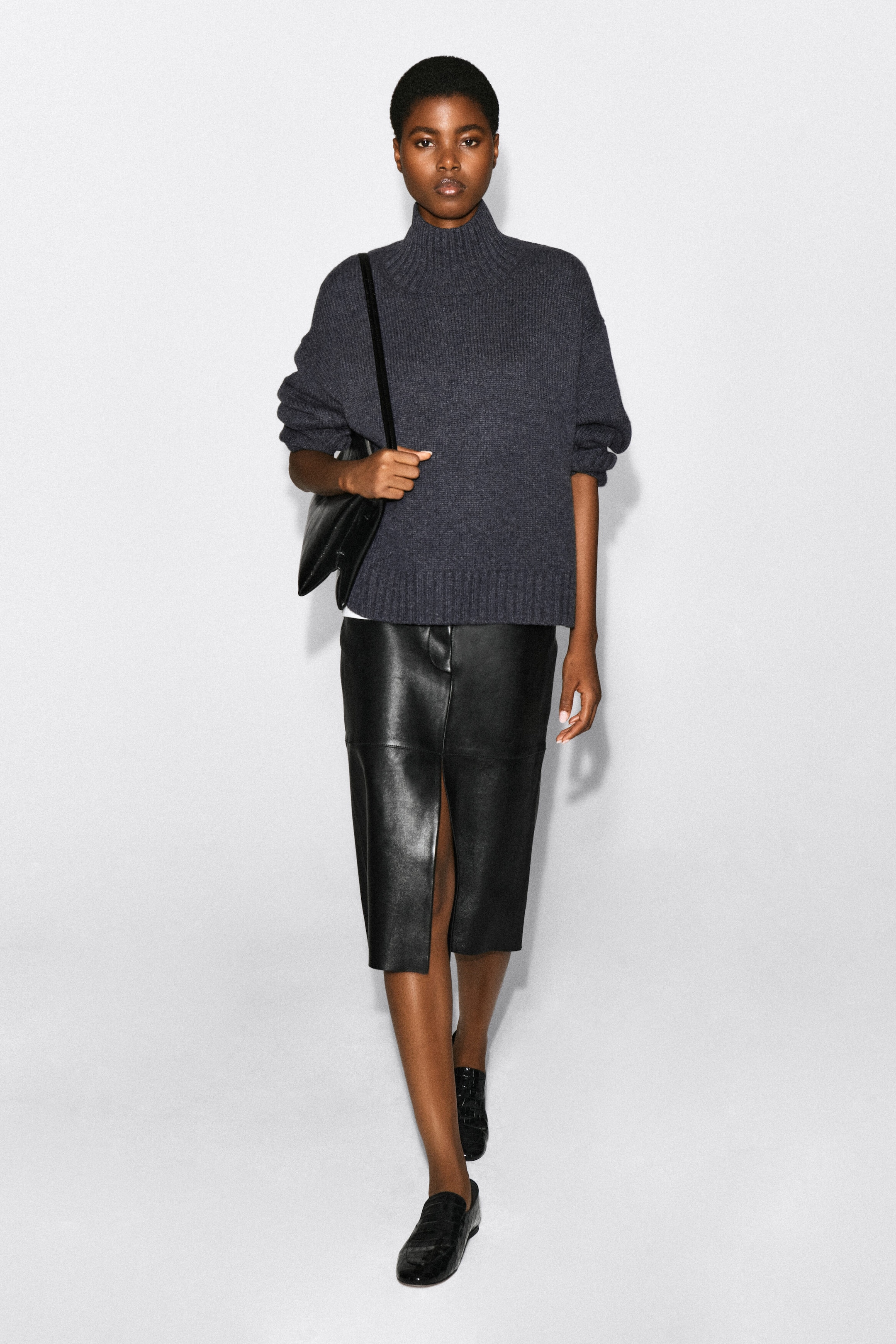A dark grey ribbed funnel-neck jumper is being worn by the model, with the sleeves slightly pushed up. The jumper is styled with a black leather midi skirt with a front slit and a black leather shoulder bag held by the strap. A dark grey ribbed funnel-neck jumper is being worn by the model, with the sleeves slightly pushed up. The jumper is styled with a black leather midi skirt with a front slit and a black leather shoulder bag held by the strap.