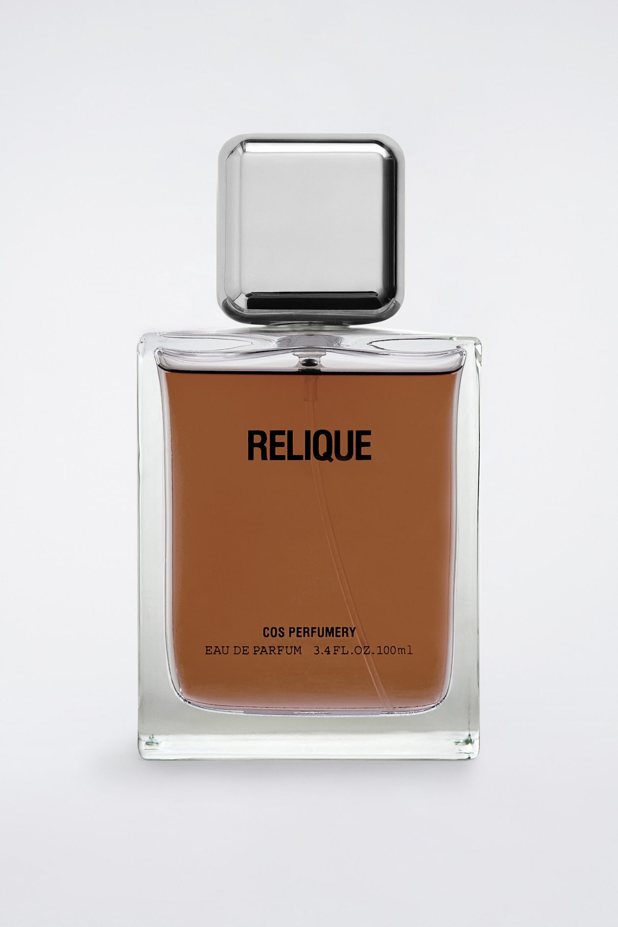 The COS Perfumery "Relique" Eau de Parfum is standing upright in a front-facing view, showcasing a rectangular clear glass bottle filled with an amber-coloured liquid. The bottle features a square, polished metallic silver cap and displays black printed text reading "RELIQUE" in the centre, with "COS PERFUMERY EAU DE PARFUM 3.4 FL.OZ. 100ml" near the base.