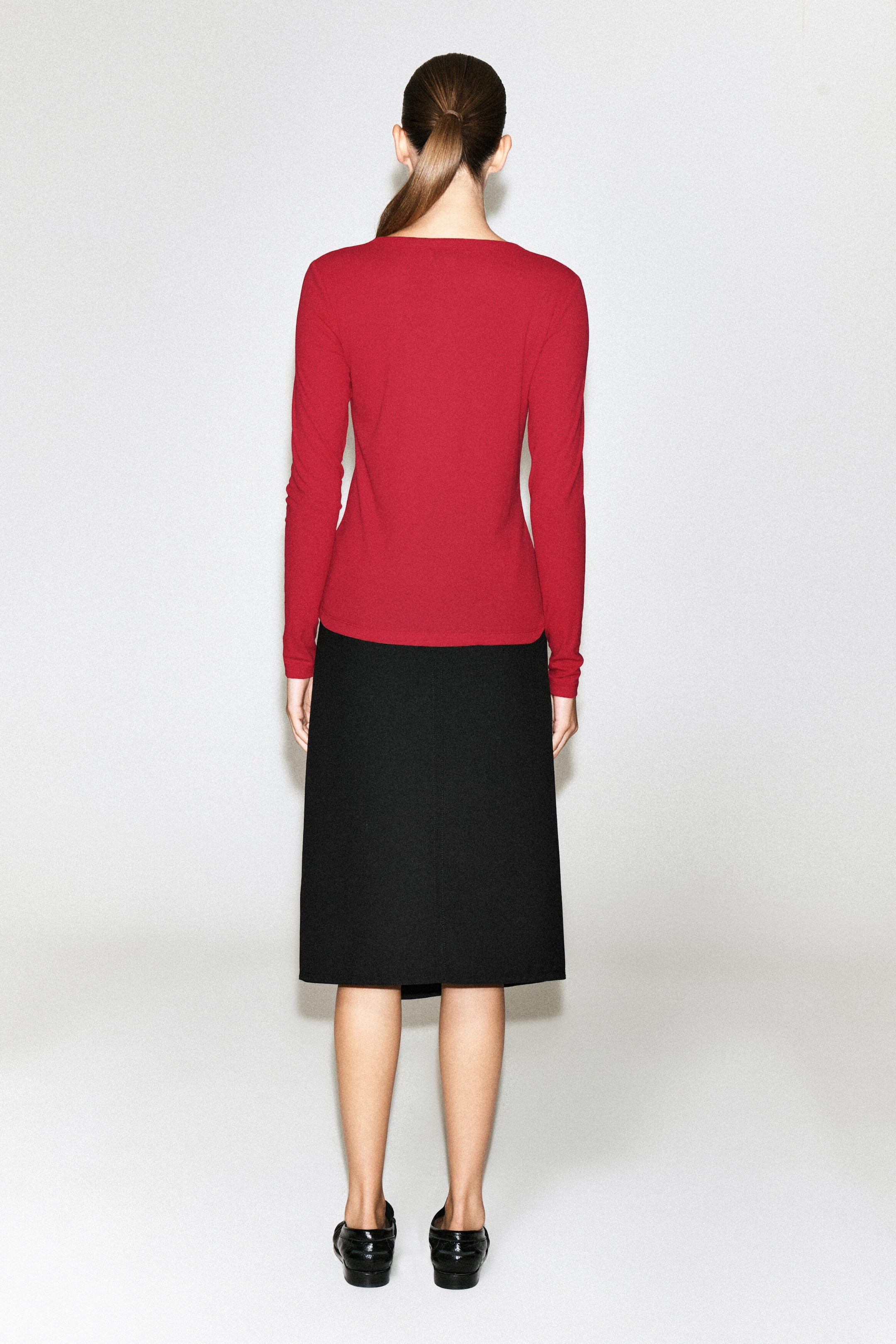 The model is wearing a red, long-sleeved jersey cardigan top, shown from the back. The slim-fitting top features a straight hem and is styled with a knee-length black skirt. The model is wearing a red, long-sleeved jersey cardigan top, shown from the back. The slim-fitting top features a straight hem and is styled with a knee-length black skirt.