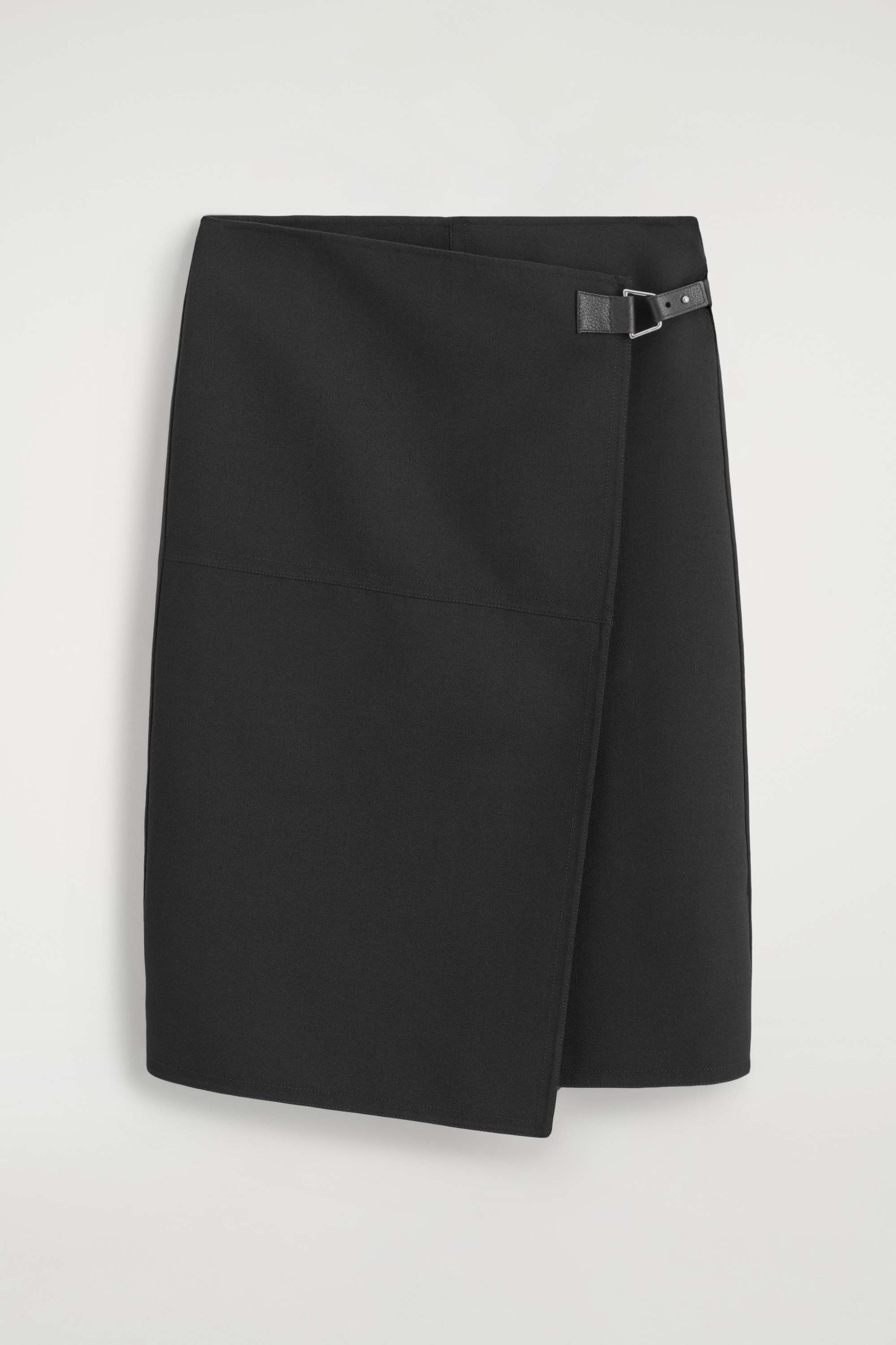 The leather-belt wool-twill midi skirt showcases a structured wrap design from the front, featuring a high-rise waist and an off-centre front slit. A small black leather belt with a buckle secures the wrap at the left hip, with a white blouse tucked in to emphasise the clean waistline.